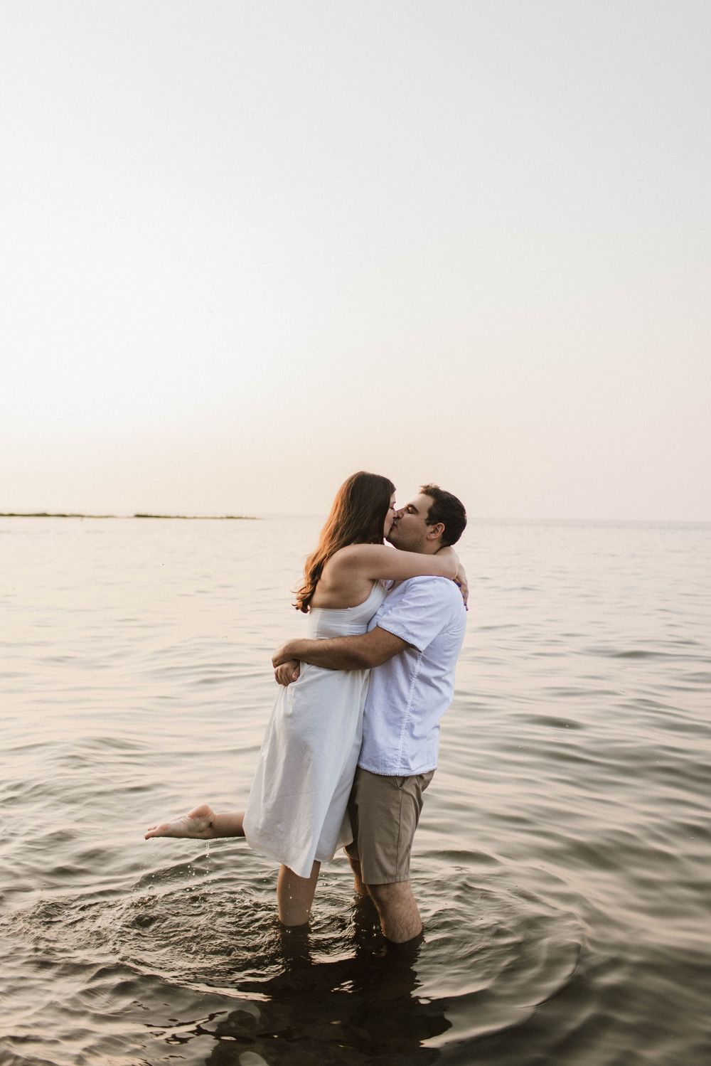 Romantic couple embracing and kissing while standing in shallow ocean water at sunset, woman in white dress.