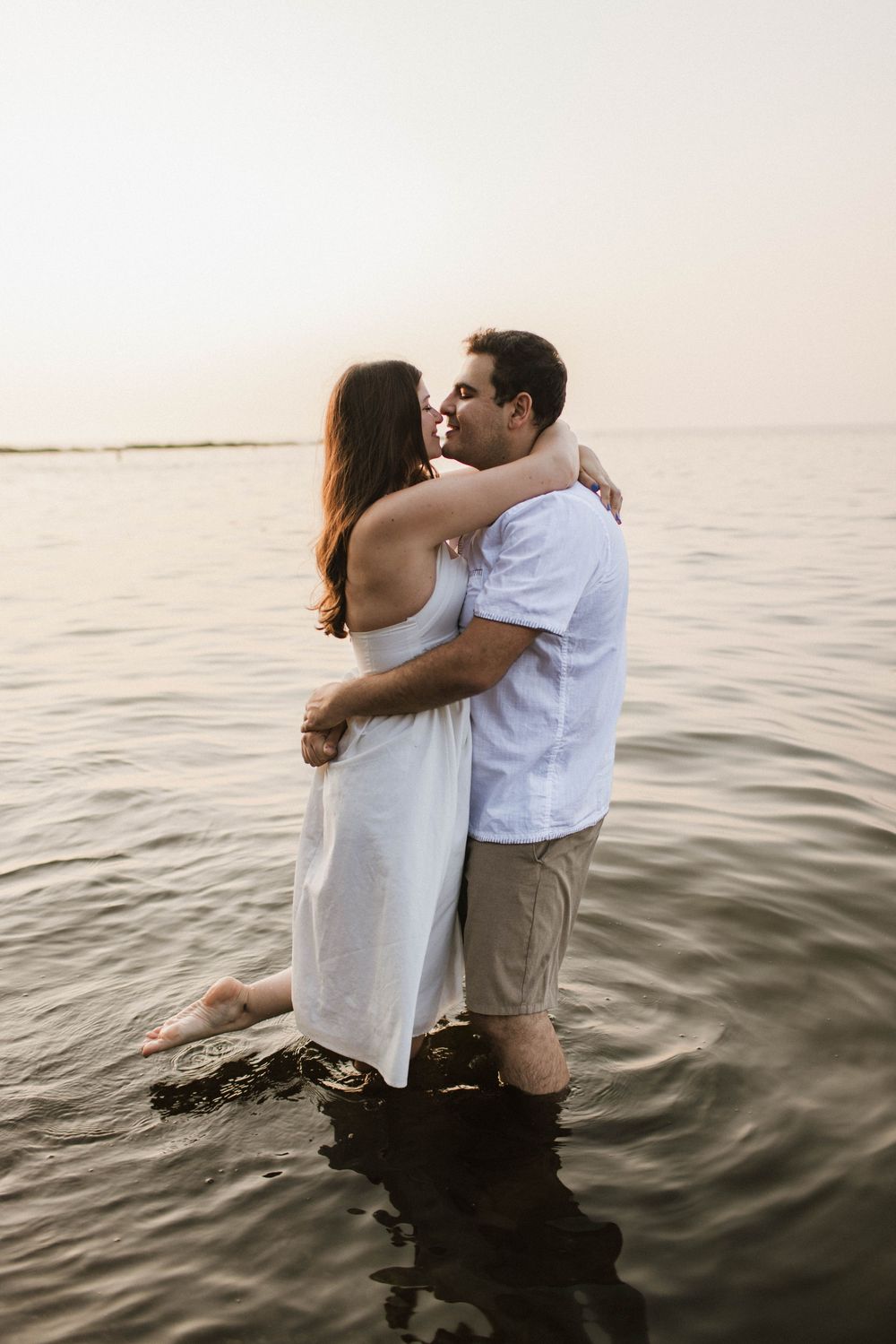 Couple sharing a tender kiss while standing in calm ocean water at dusk, woman in white dress, man in blue shirt.