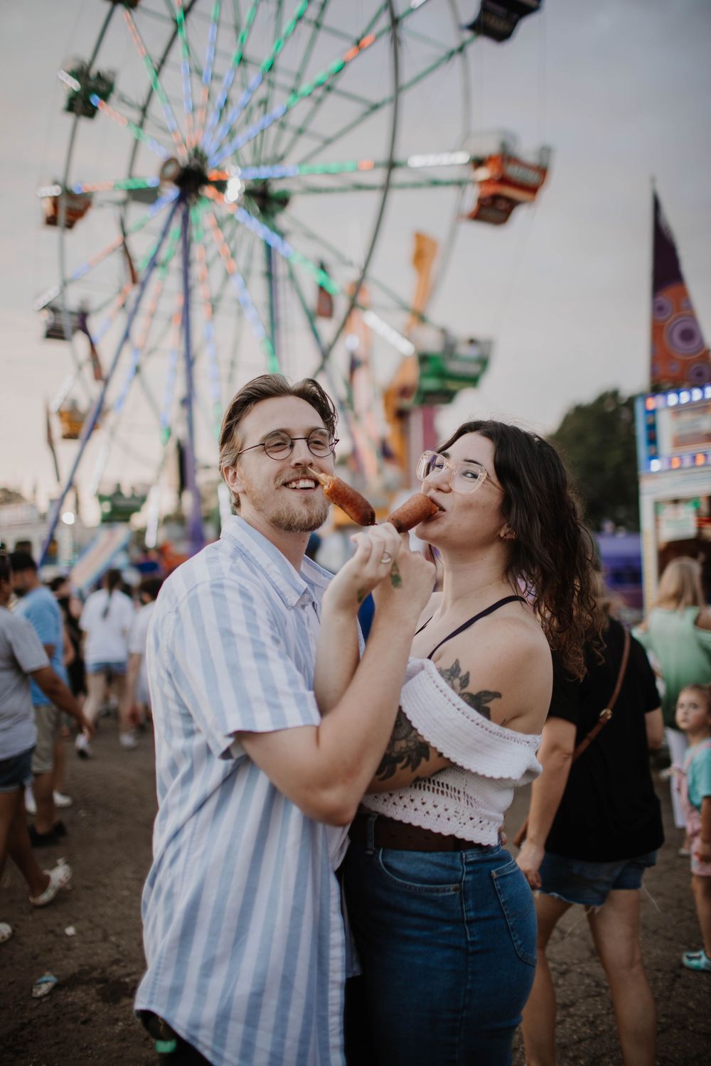 Couple sharing a corn dog at a summer fair with a Ferris wheel and colorful lights in the background.