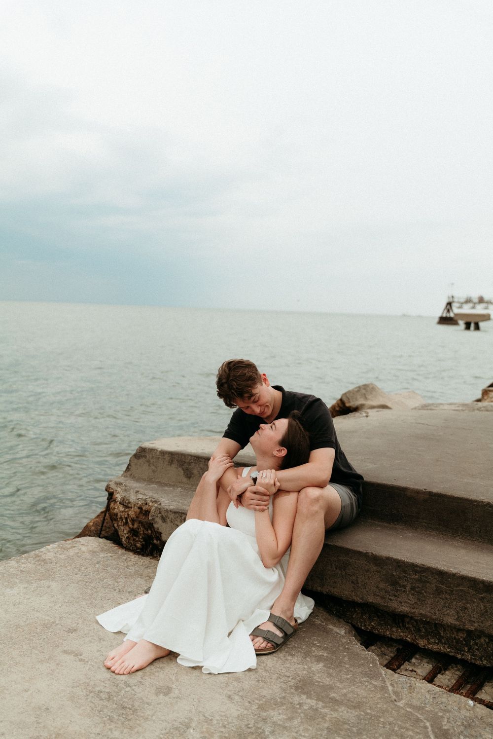 Couple sharing a romantic kiss on seaside stone steps, woman in white dress, ocean backdrop.