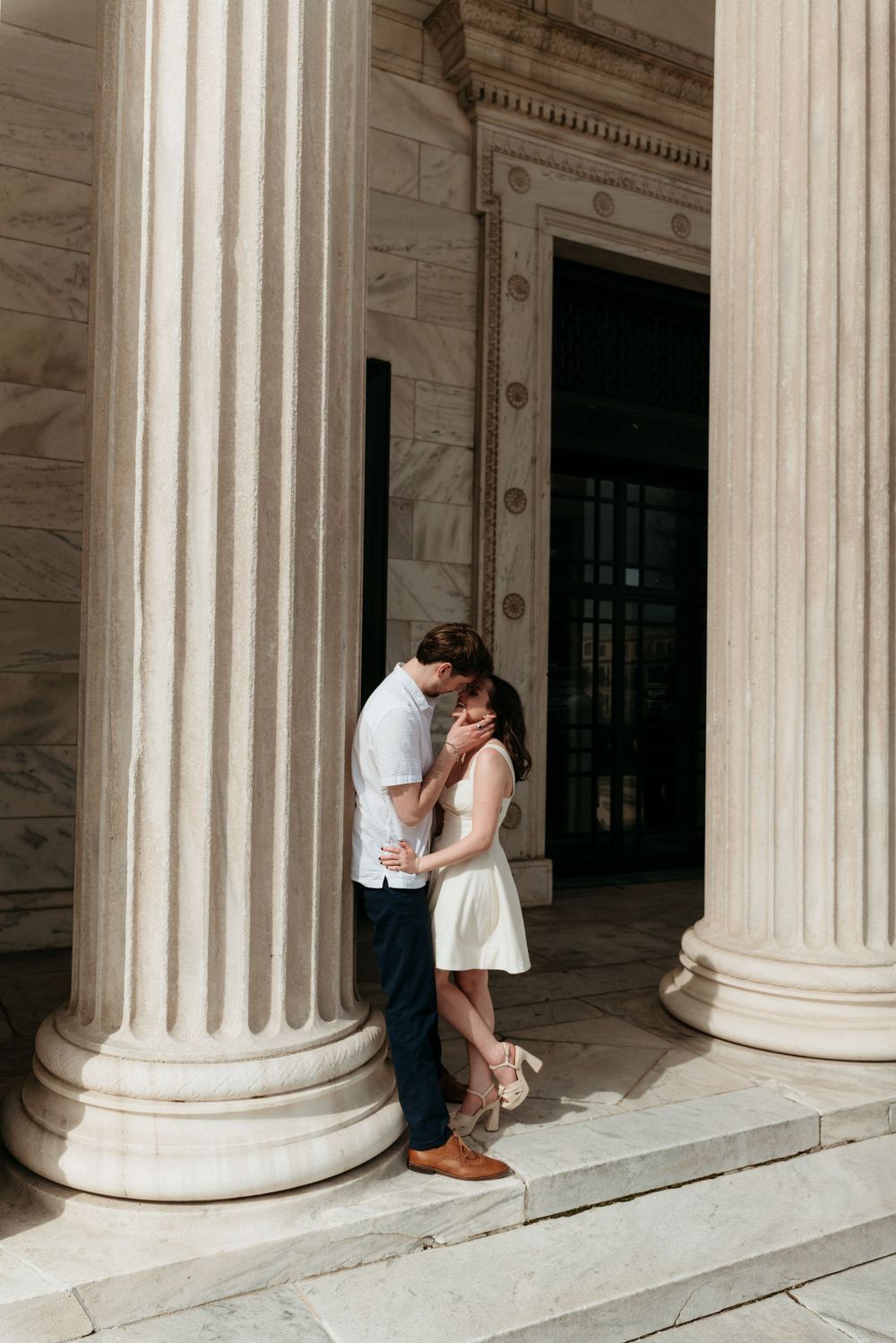 Couple sharing a romantic kiss between grand marble columns of a neoclassical building on sunlit steps.