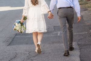 Bride in white lace dress holding blue bouquet walks hand-in-hand with groom in gray pants on pavement.