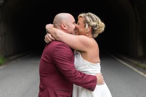 Bride and groom sharing a romantic kiss in front of a dark tunnel, groom in burgundy suit, bride in white dress.