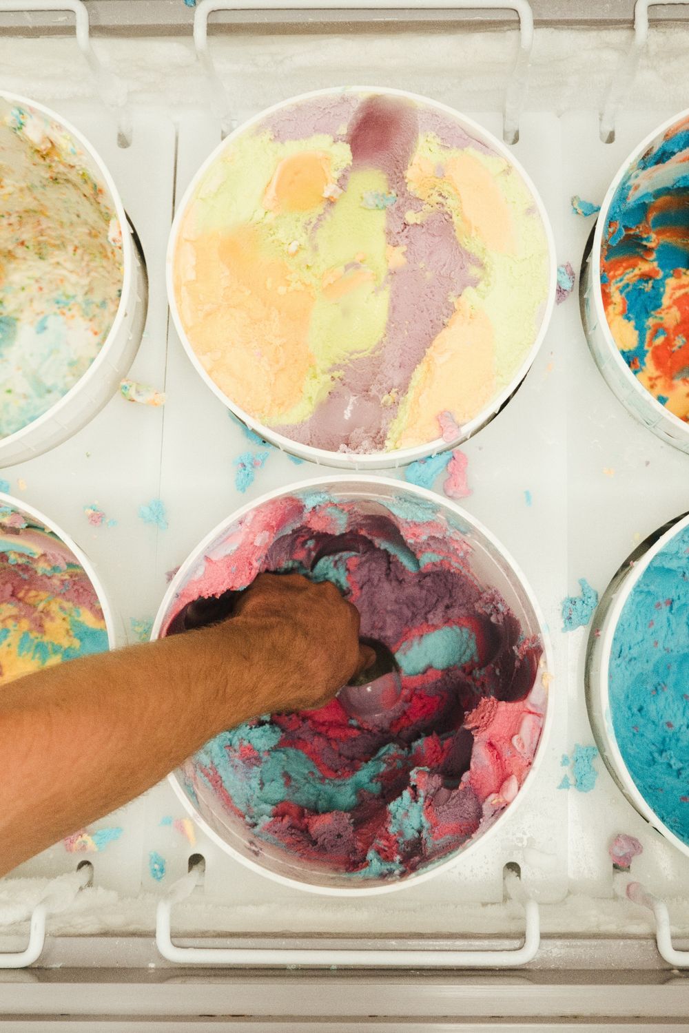 Hand stirring vibrant multicolored ice cream in metal containers at a gelato shop display case.