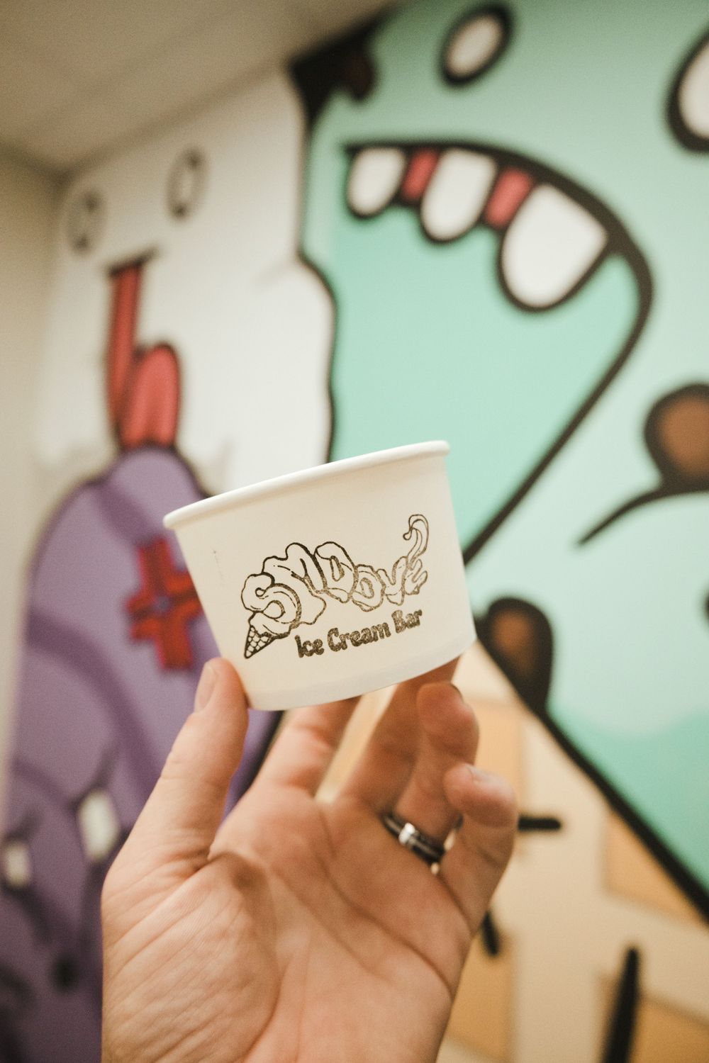 Hand holding a Sno-Biz Ice Cream Bar paper cup in front of colorful graffiti mural.