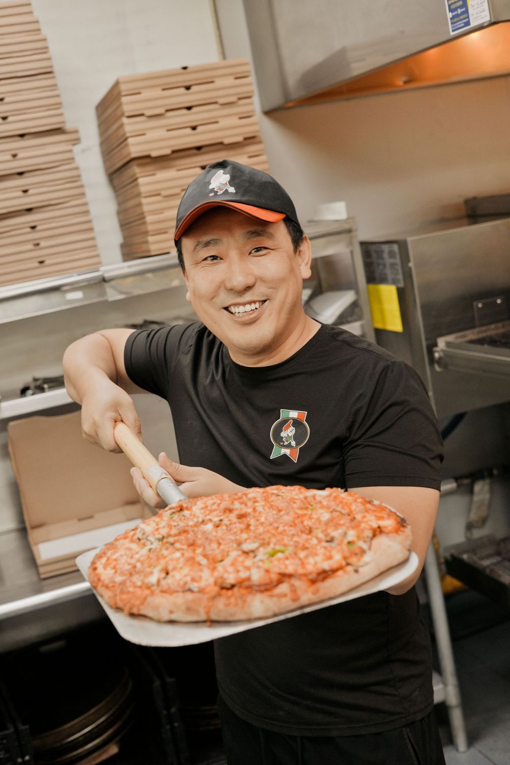 Smiling man in black cap holds a large pepperoni pizza on a peel in a pizzeria kitchen.