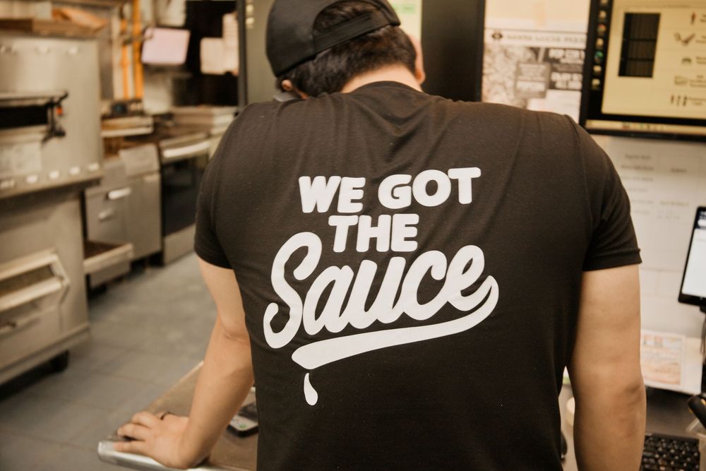 Person wearing a black 'We Got The Sauce' t-shirt working in a commercial kitchen.