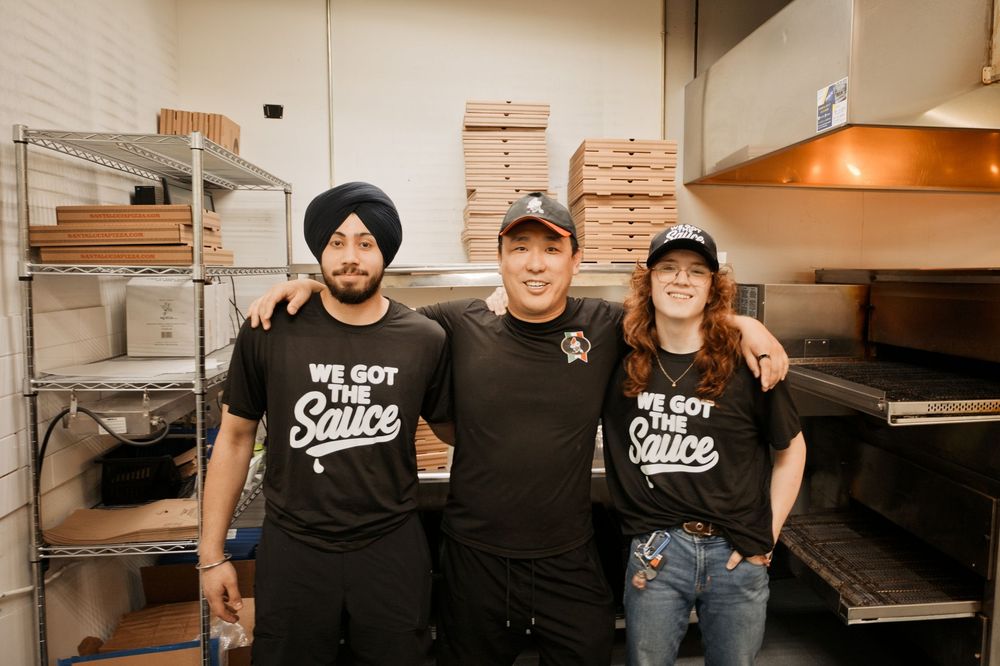 Three restaurant staff members in black We Got The Sauce shirts smile together in a commercial kitchen.