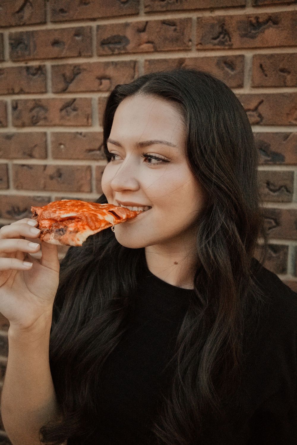 Young woman with long dark hair eating a slice of pizza against a brick wall background.