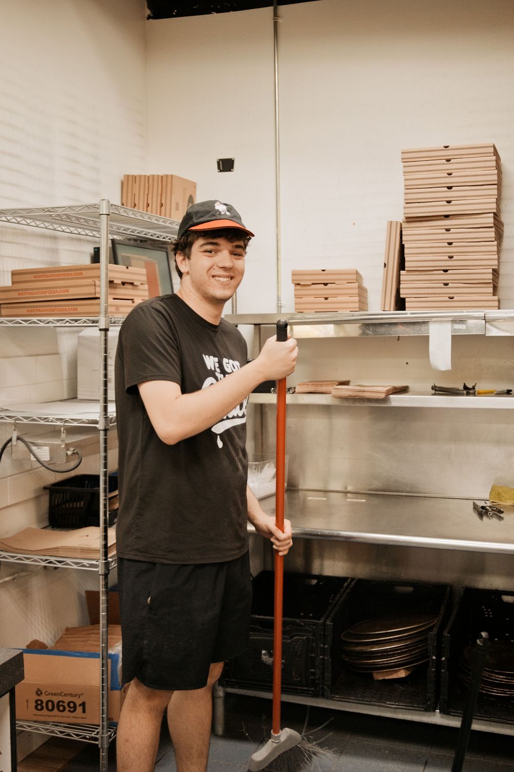Young man in black t-shirt and cap smiling while sweeping a restaurant storage room with pizza boxes on shelves.