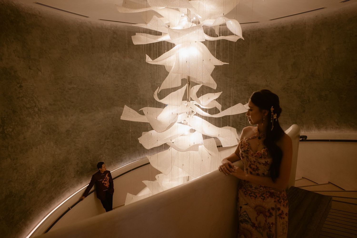 Woman in floral dress standing on spiral staircase beneath artistic paper chandelier in dimly lit venue.