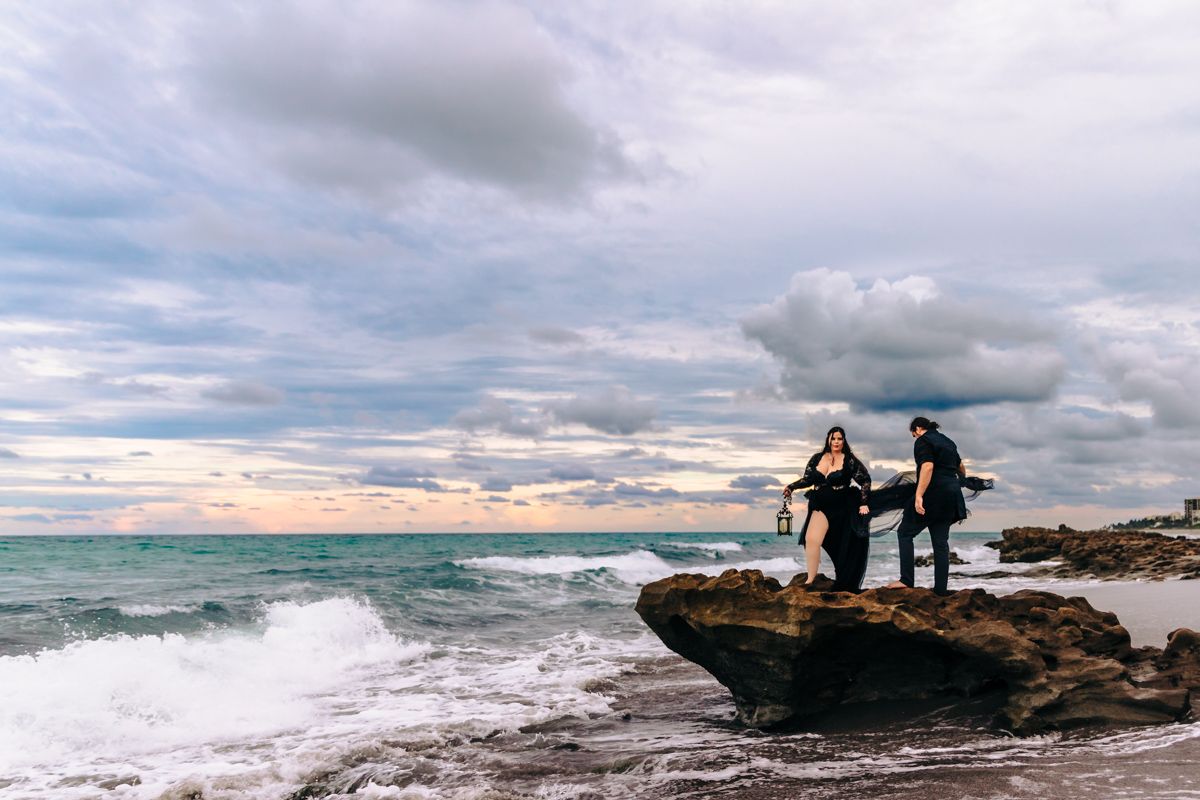Couple standing on rocky Jupiter beach shore with crashing waves and dramatic cloudy sky at sunset on blowing rocks beach in Jupiter.