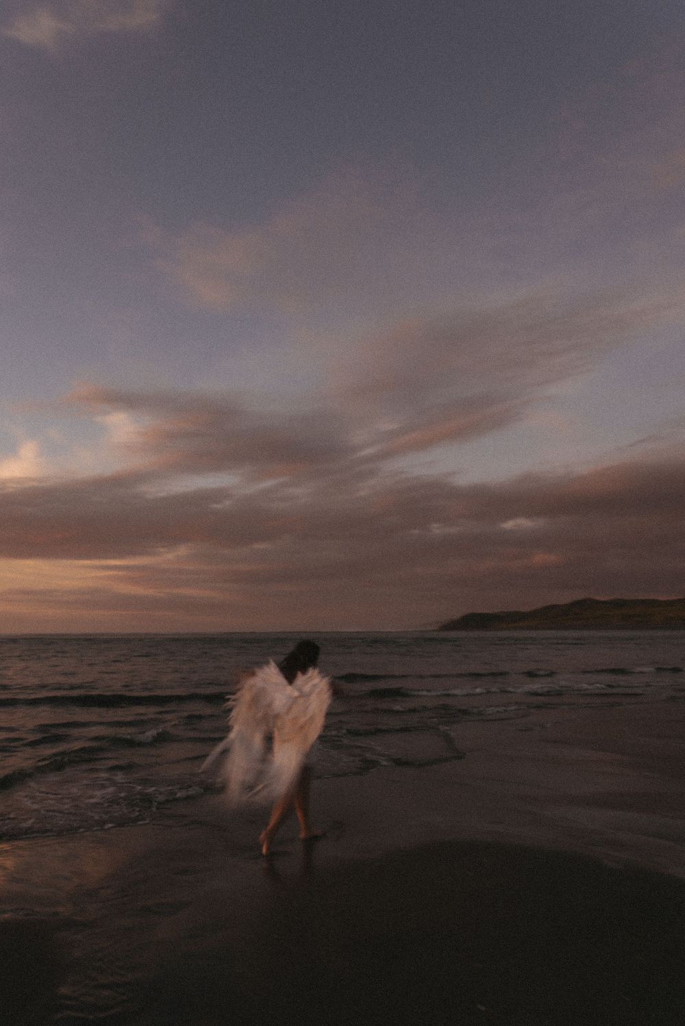 A person wrapped in white feathers stands on a beach at sunset with dramatic clouds and ocean waves.