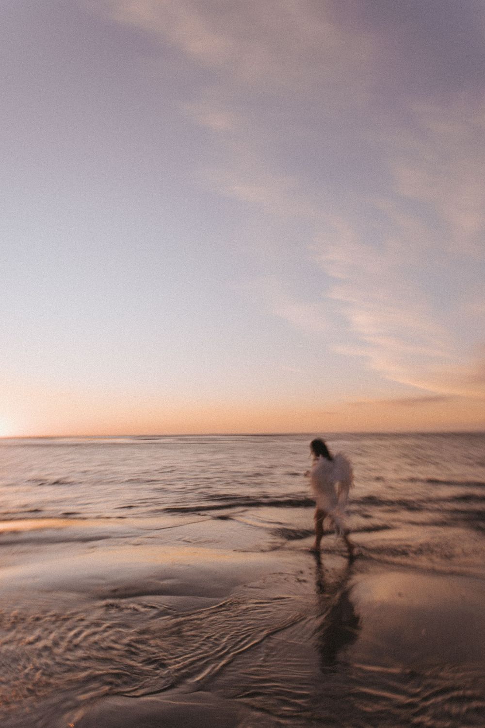 A lone figure in white walks along a serene beach shore at sunset, with soft waves and pastel skies.