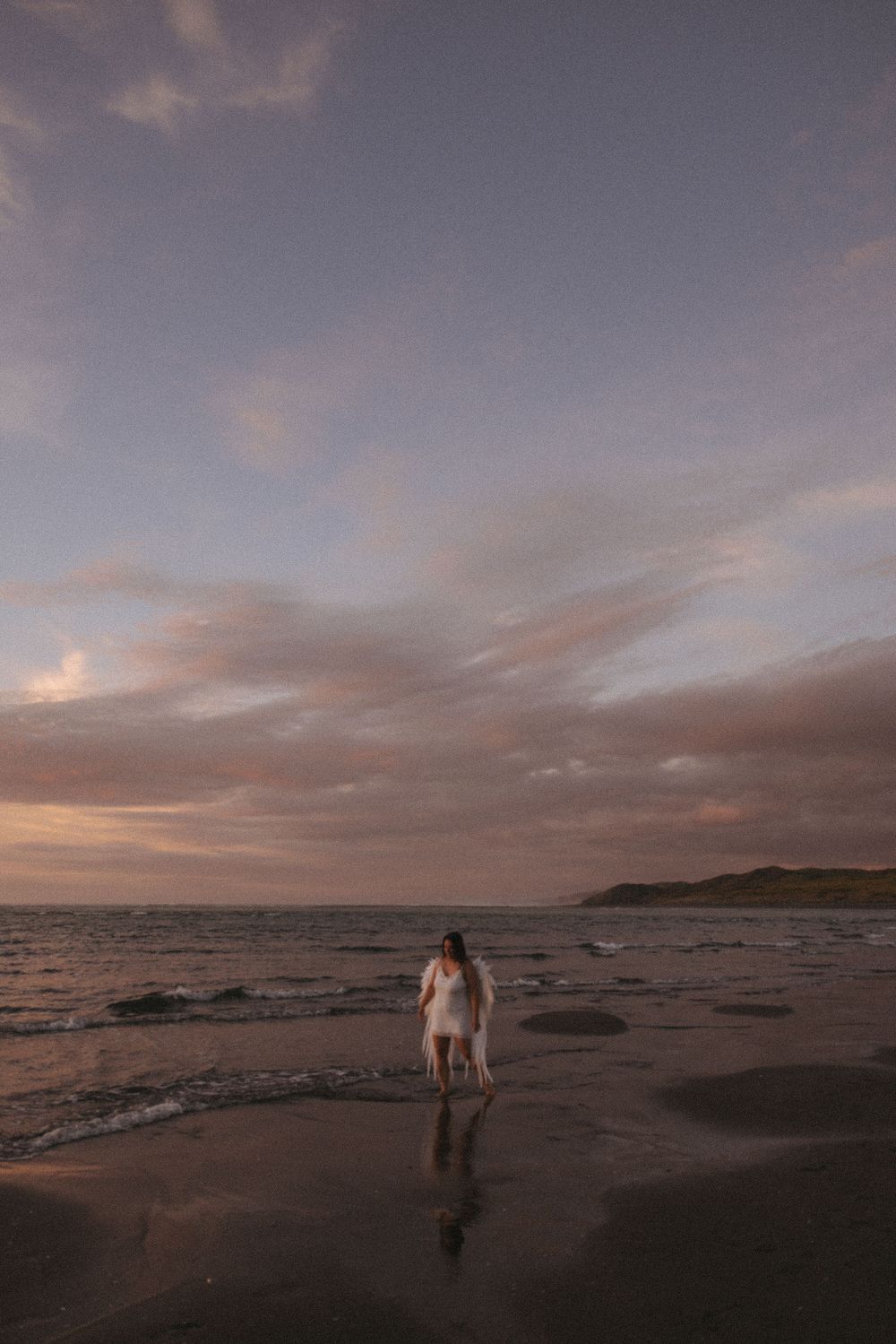 Woman standing on a reflective beach at sunset with pink clouds and ocean waves in the background.