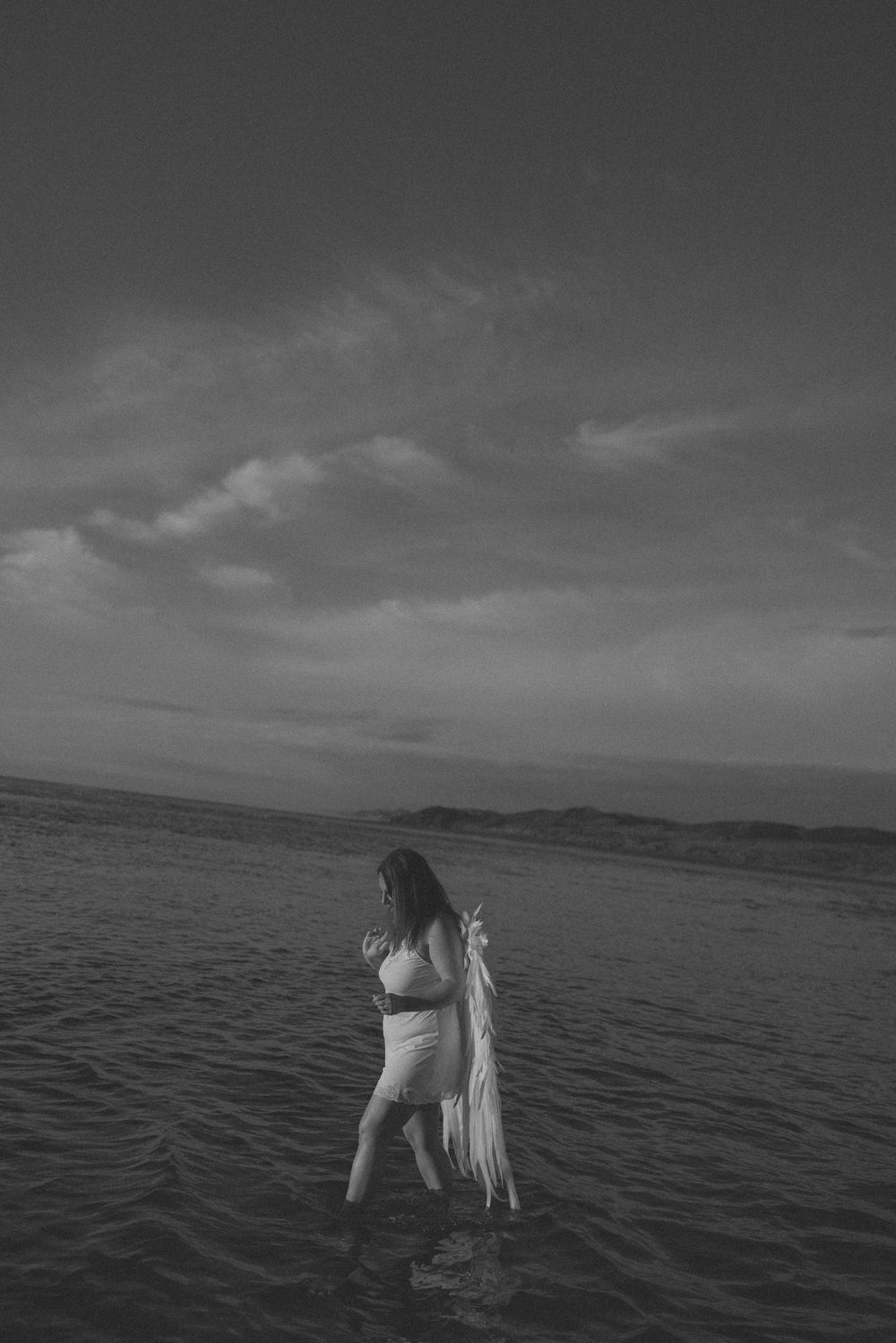 Black and white photo of a woman standing in ocean water holding a white fabric under a dramatic cloudy sky.