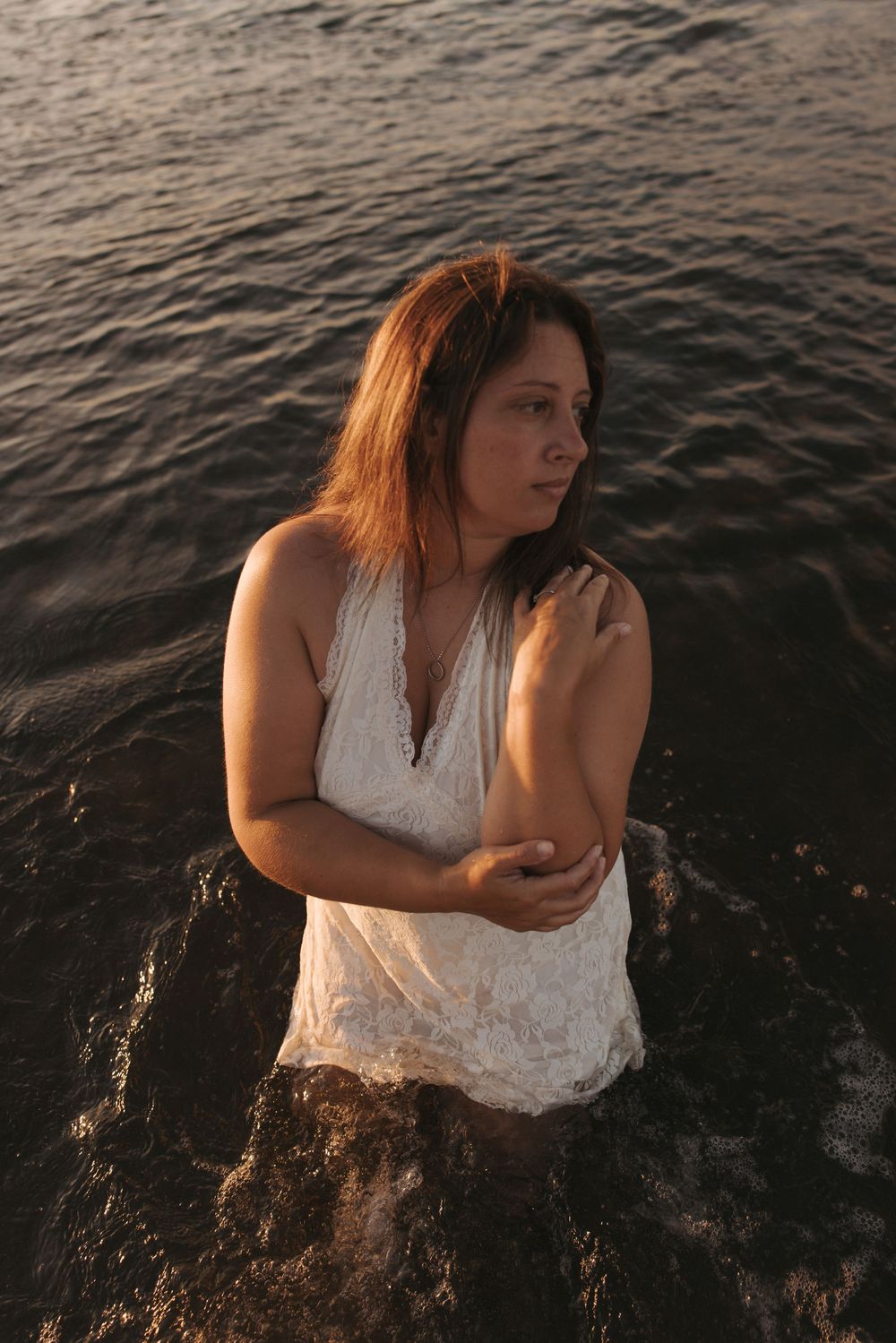 Woman in white lace dress standing in ocean waves at golden hour sunset, looking away thoughtfully.