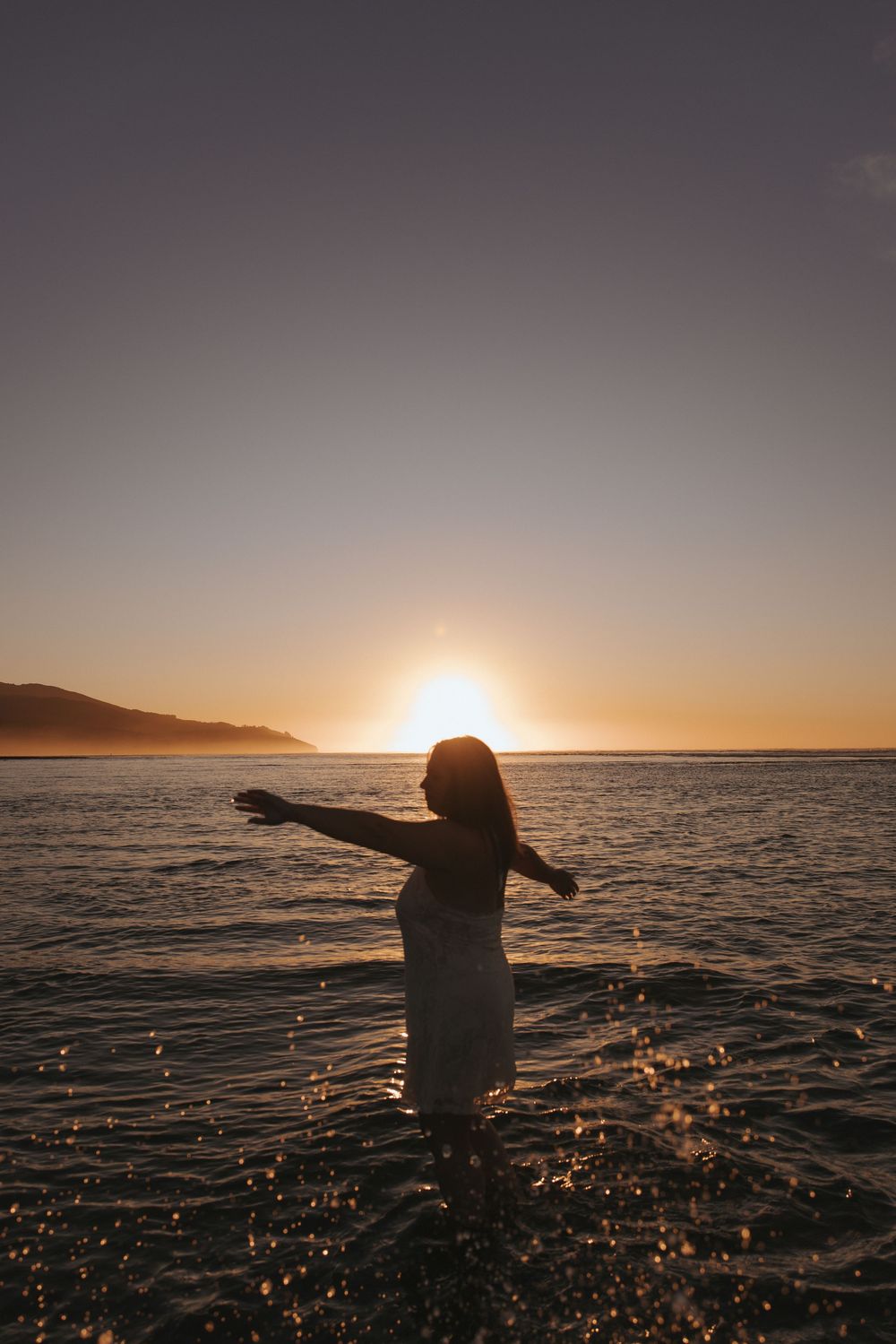 Silhouette of a woman standing in the ocean with arms outstretched at golden sunset with mountains in background.