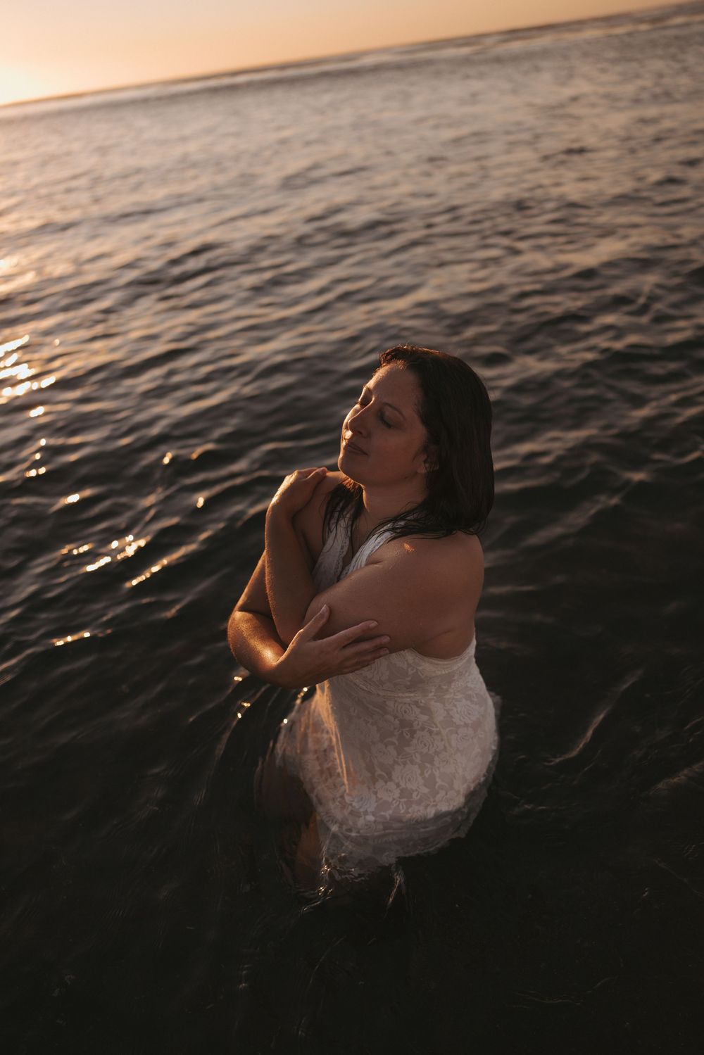 Woman in white dress standing in ocean water at sunset, eyes closed, hands on shoulders.