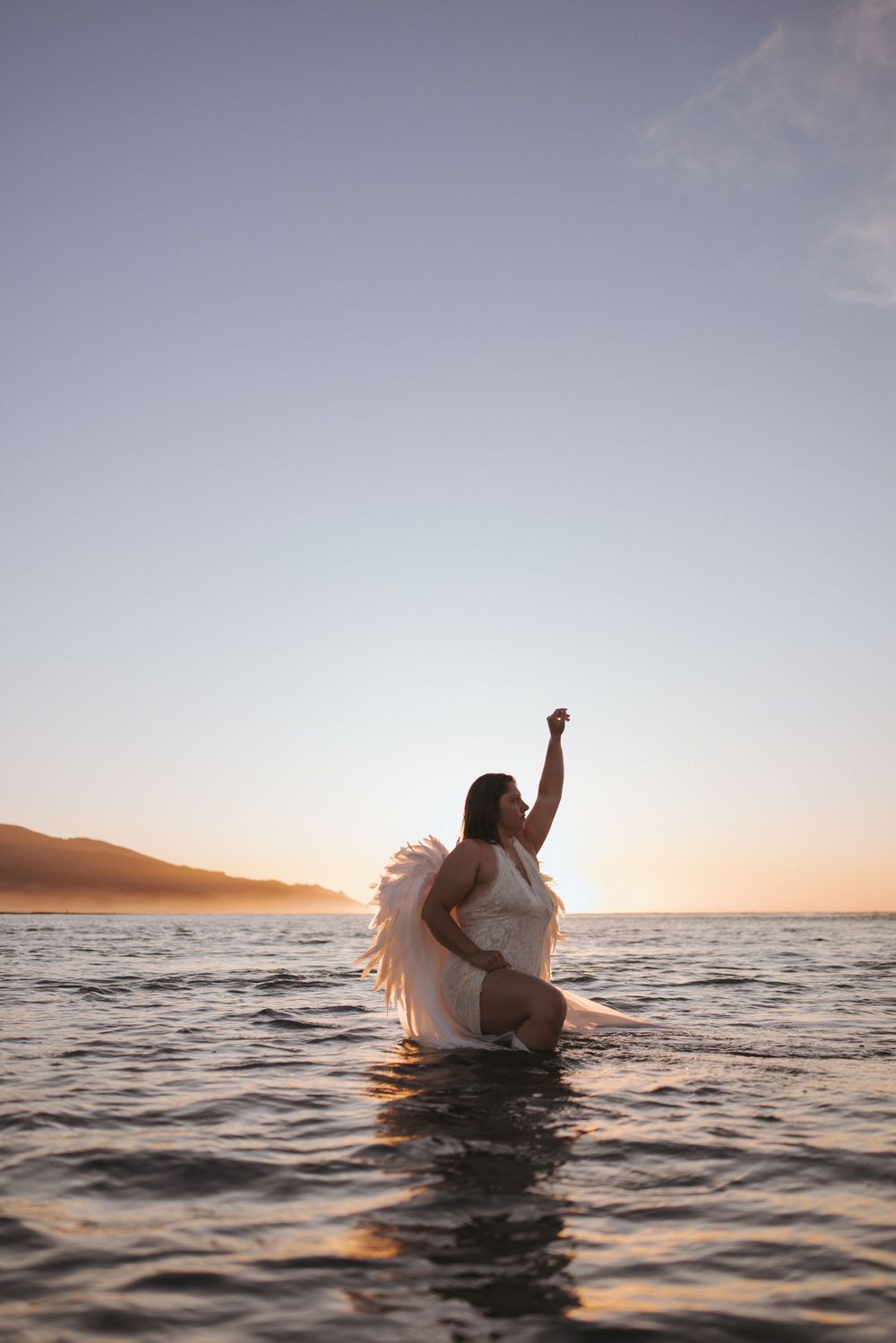 Woman in white dress with angel wings stands in ocean water, arm raised toward sky at golden sunset.