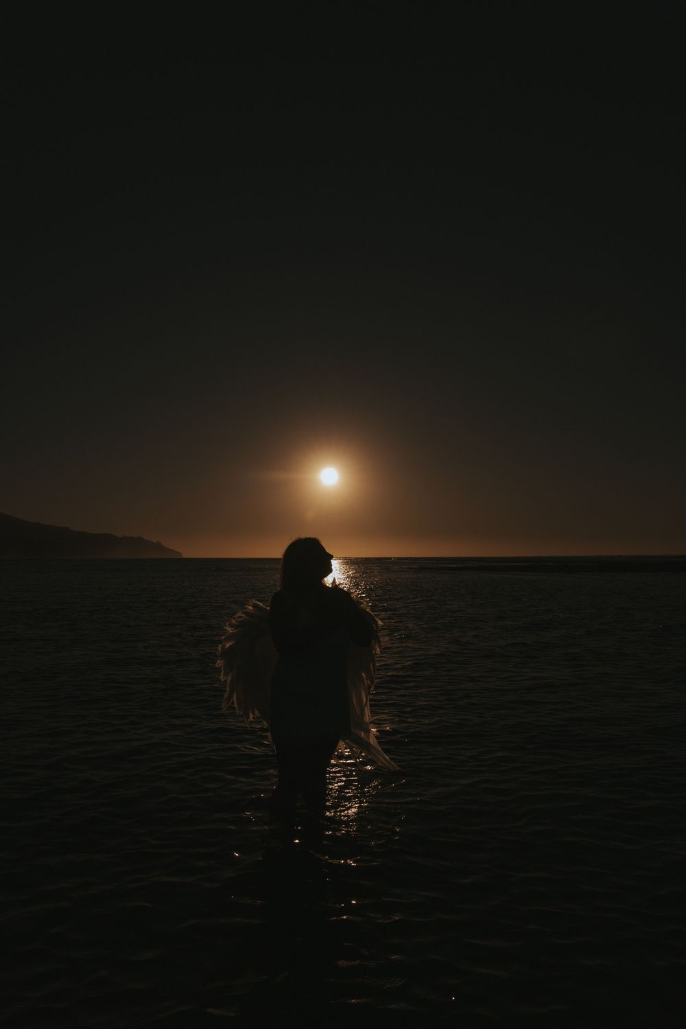 Silhouette of a person standing in moonlit ocean water at night with glowing full moon reflecting on the sea.