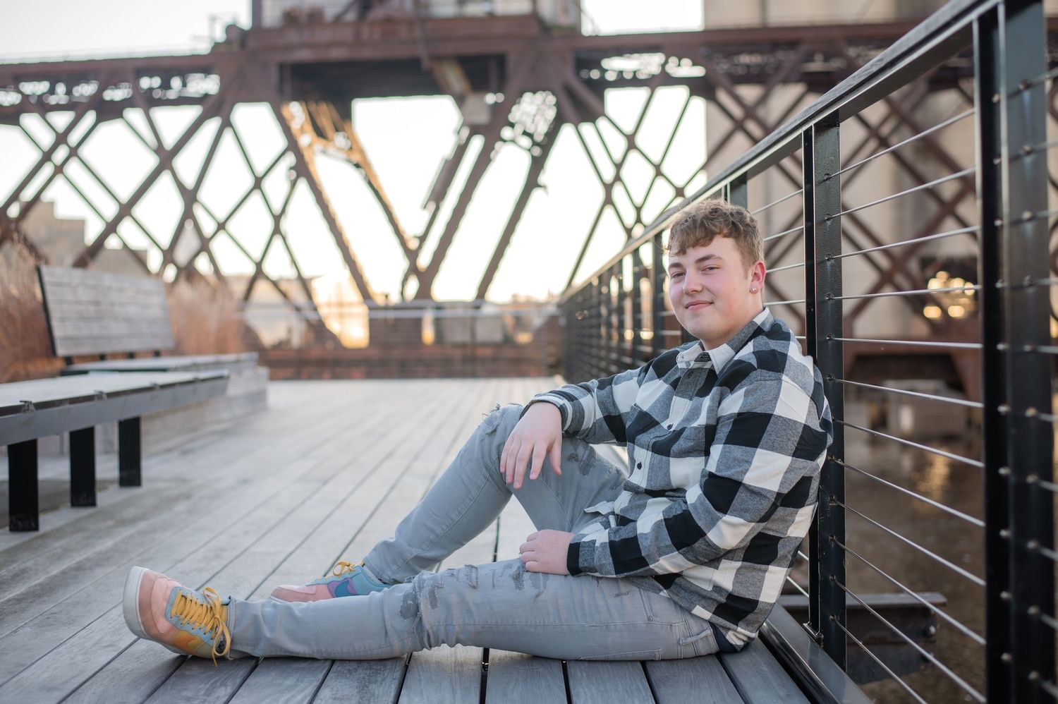 Young man in plaid shirt and jeans sitting on wooden deck near industrial bridge at sunset.