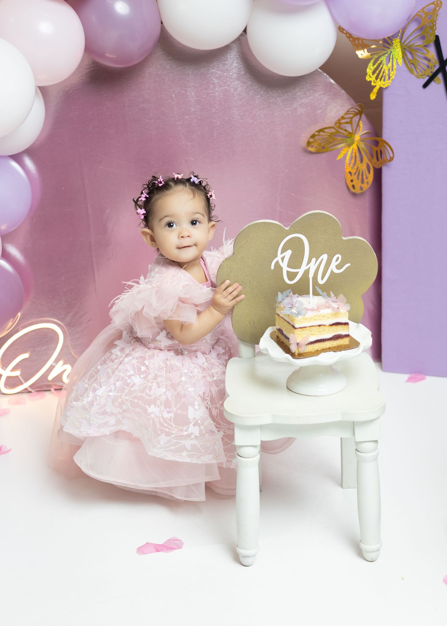 A baby girl in a pink dress celebrates her first birthday with a cake smash setup featuring balloons and butterfly decorations.