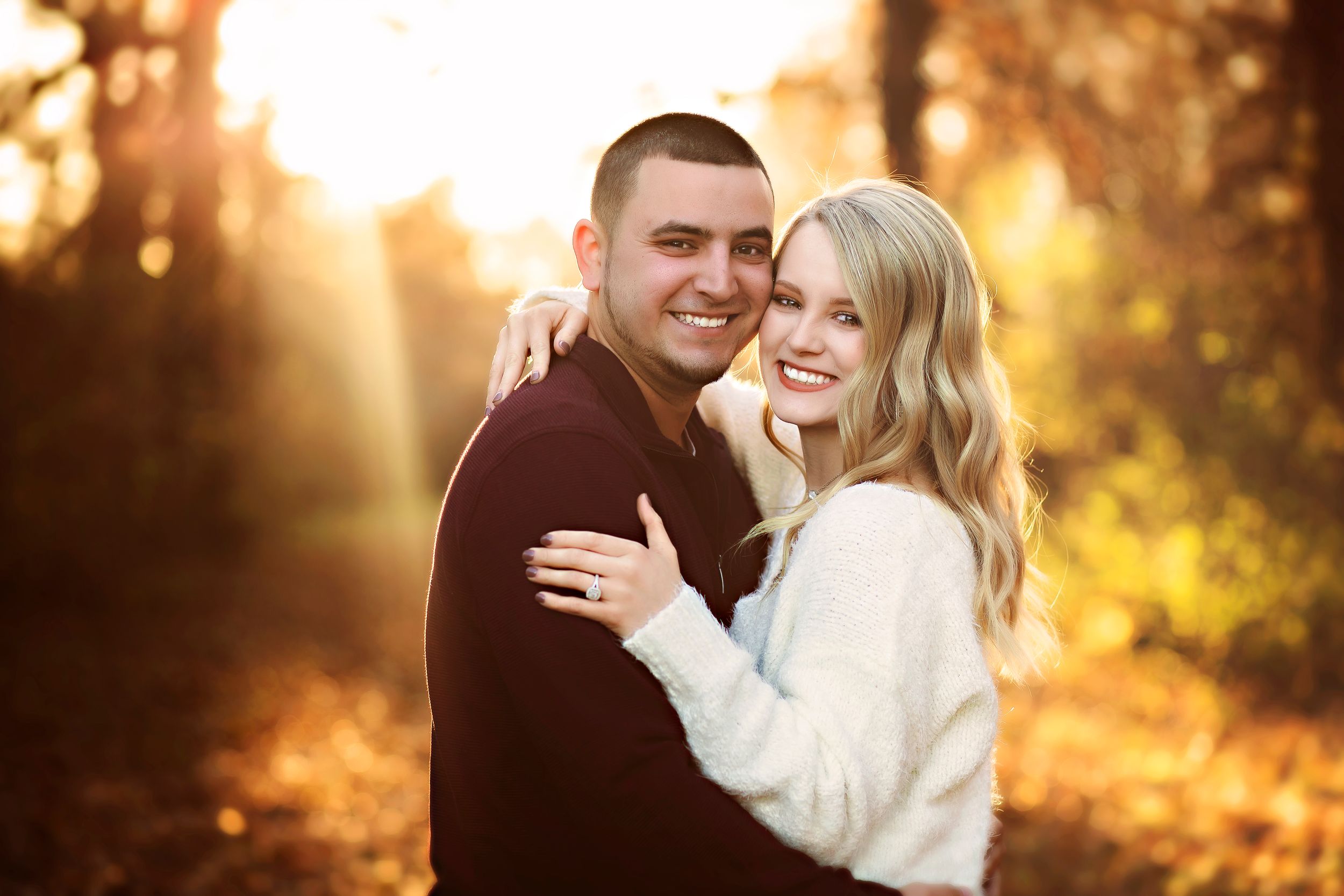 Couple embracing outdoors in golden autumn light, smiling warmly among fall foliage for an engagement photo.