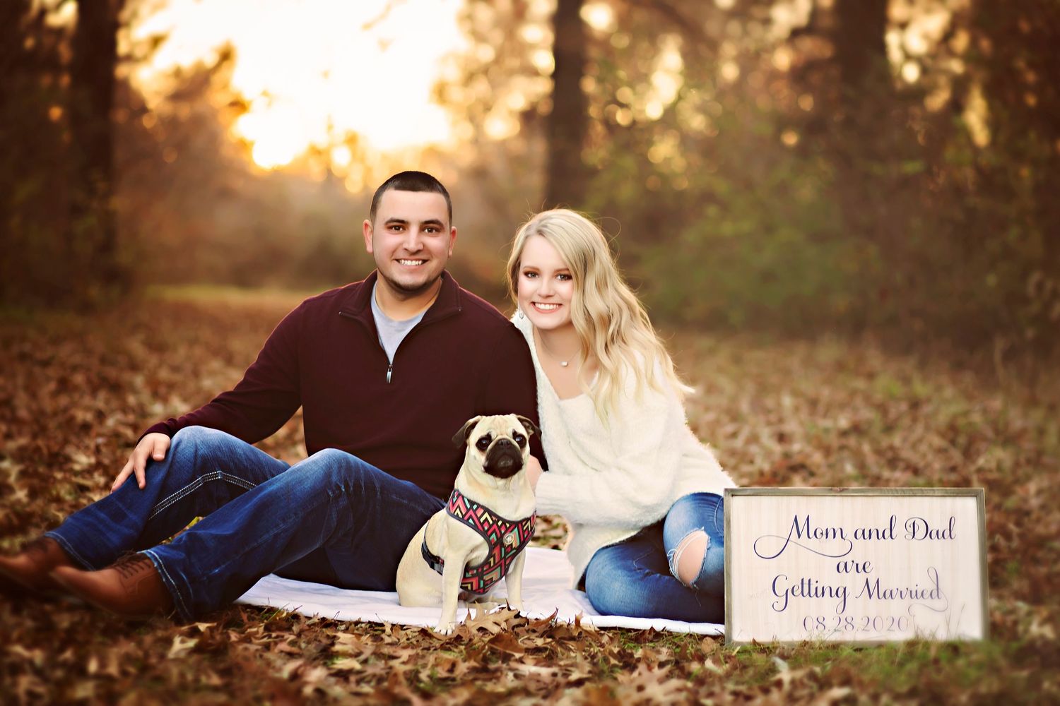 Couple sitting outdoors with pug dog holding engagement sign among autumn leaves in golden hour light.