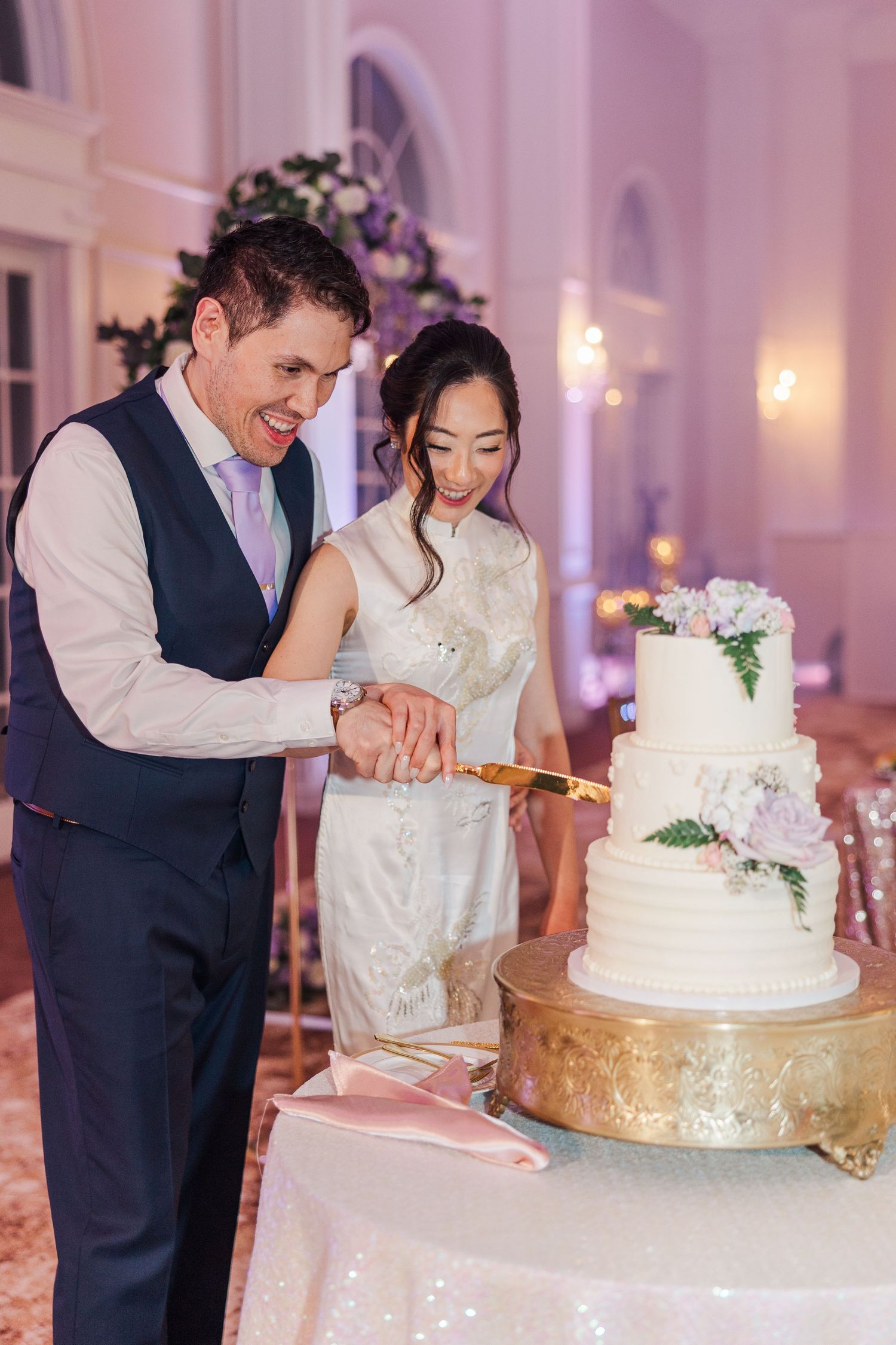 Bride and groom cutting white four-tier wedding cake decorated with flowers at elegant reception venue.