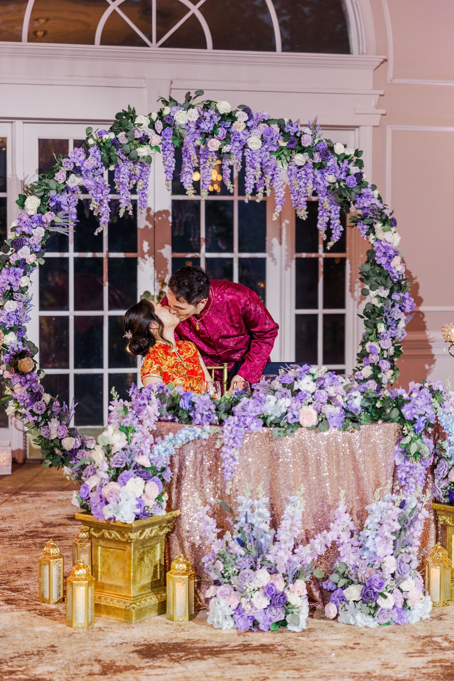 Couple kissing at sweetheart table adorned with purple floral arch and hydrangeas at elegant wedding reception.