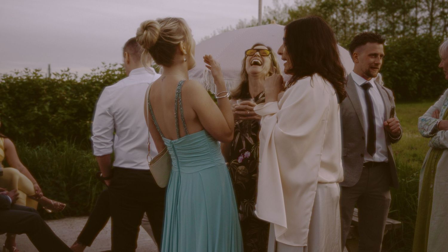 Guests in elegant attire socializing outdoors at a wedding reception, with a woman in a teal gown laughing joyfully.