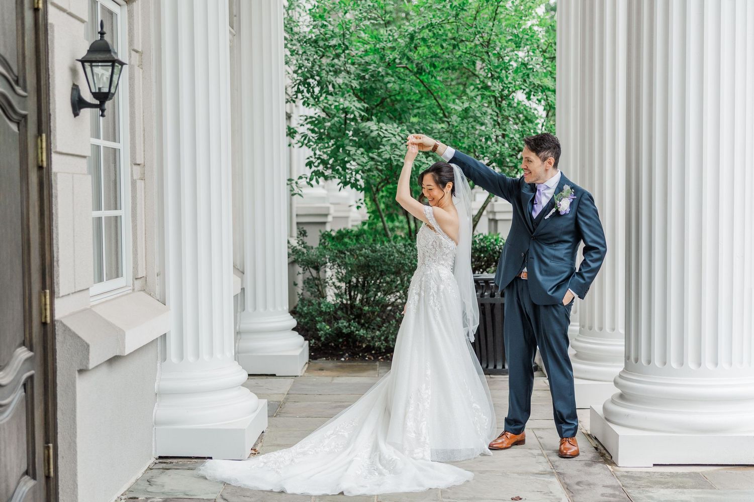 A bride and groom dance elegantly between white columns at a classic wedding venue surrounded by lush greenery.