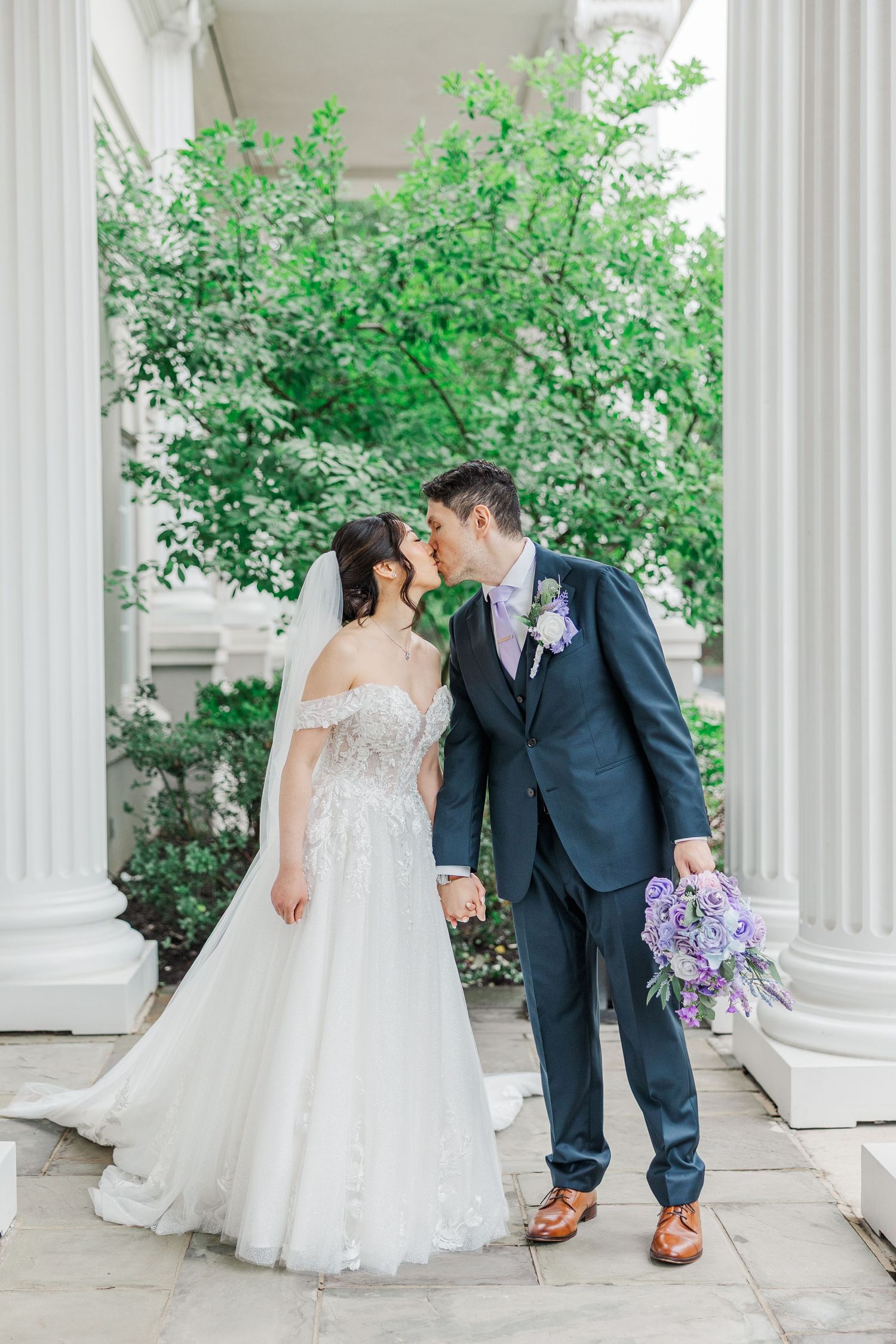 Bride and groom share a kiss between white columns with lush green trees, bride holding purple bouquet in white lace gown.