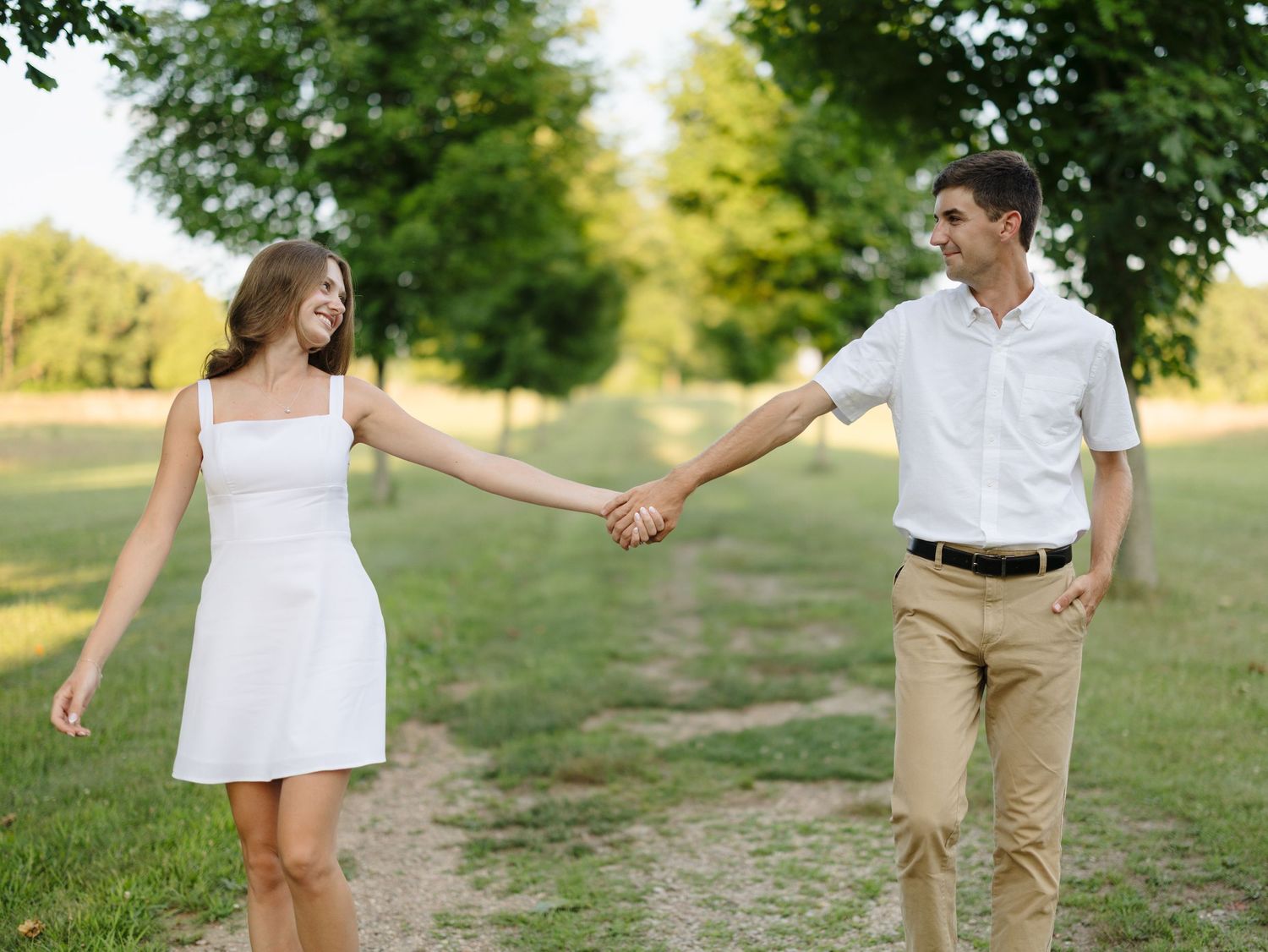 Happy couple holding hands while walking on a scenic park path surrounded by lush green trees.