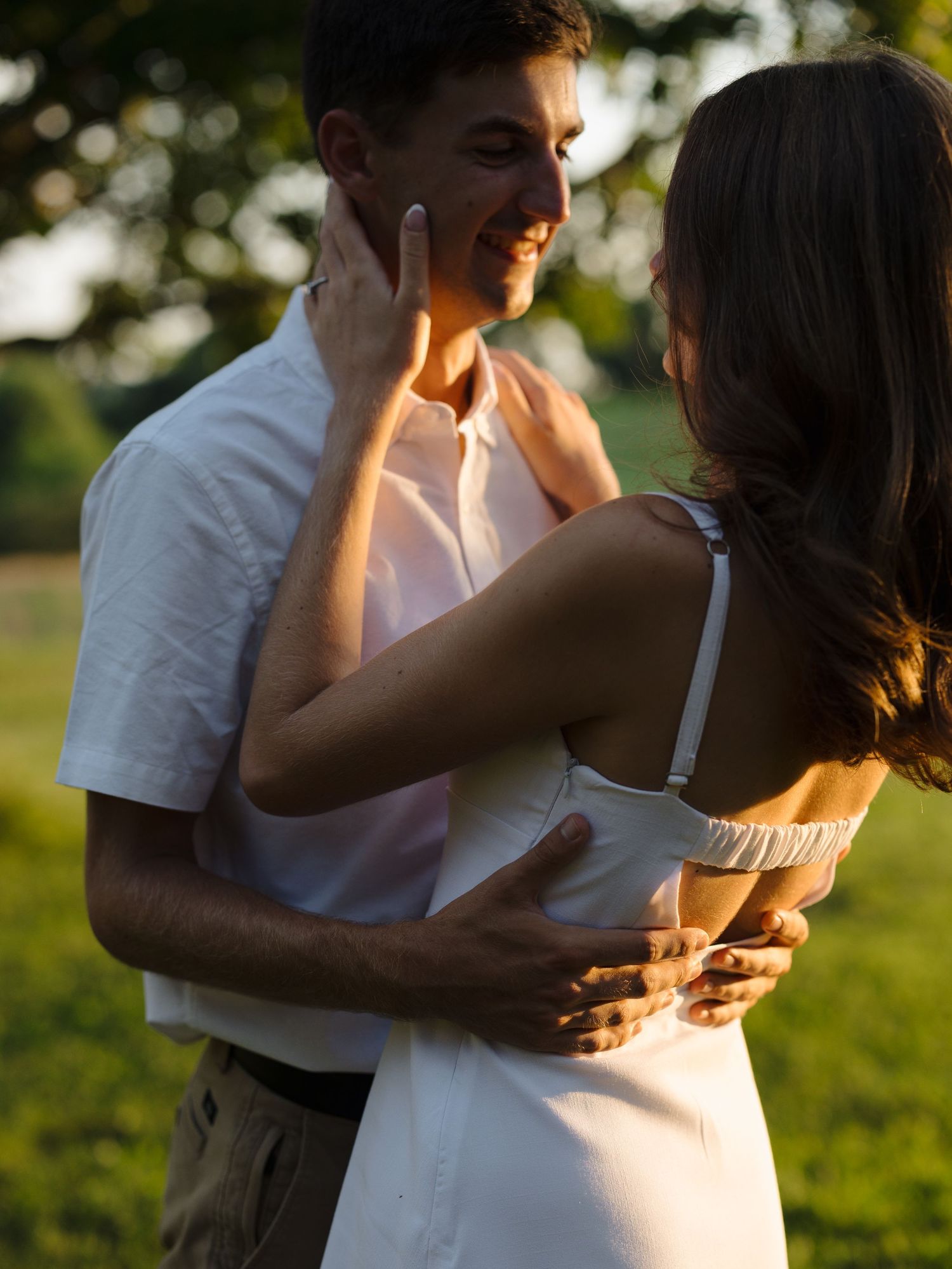 A couple in white outfits share a romantic embrace outdoors at golden hour, with the woman touching the man's face.