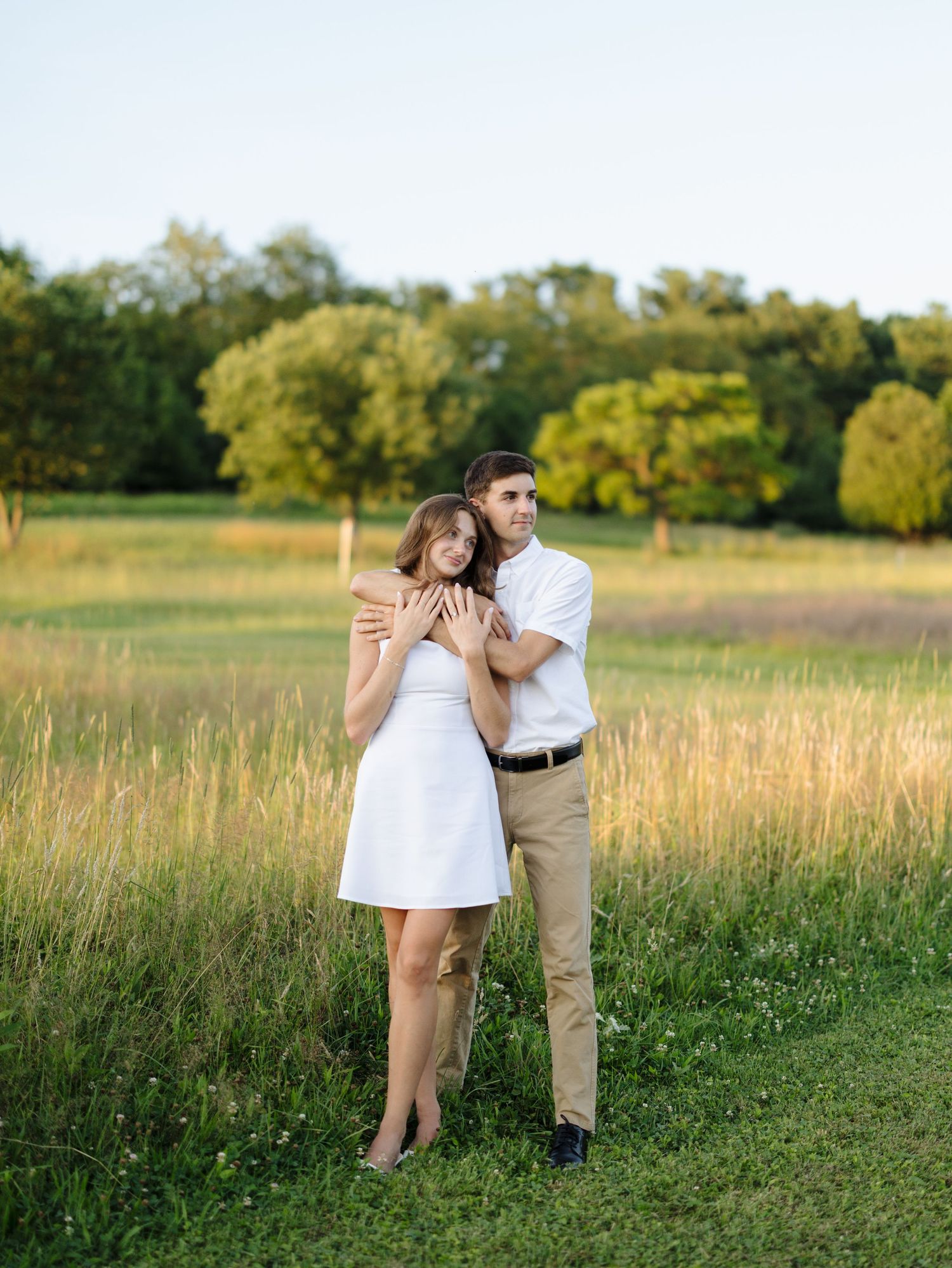 Man hugging woman from behind in a golden sunlit grassy field at sunset.