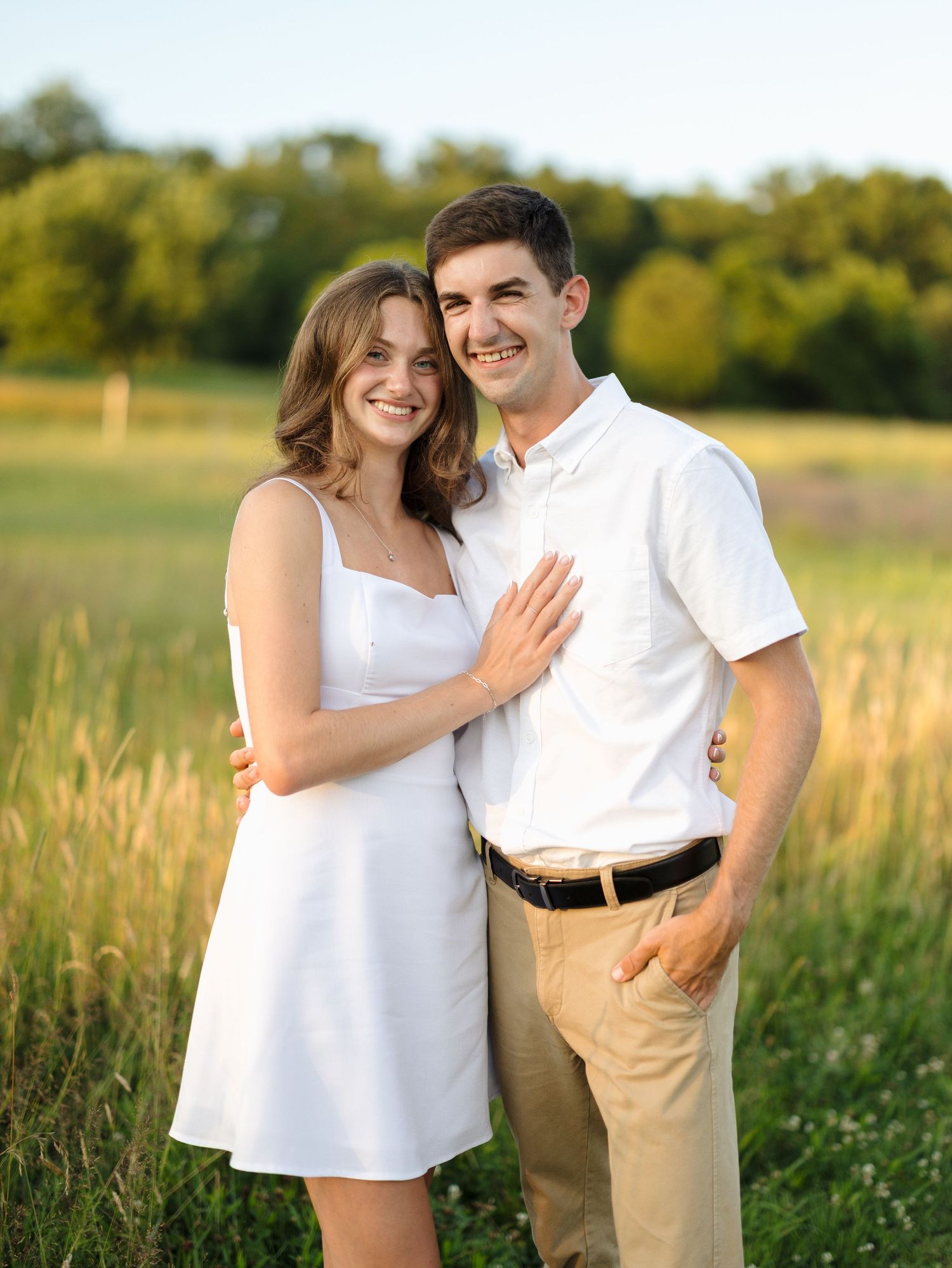 Smiling engaged couple posing together in a lush green meadow during golden hour.