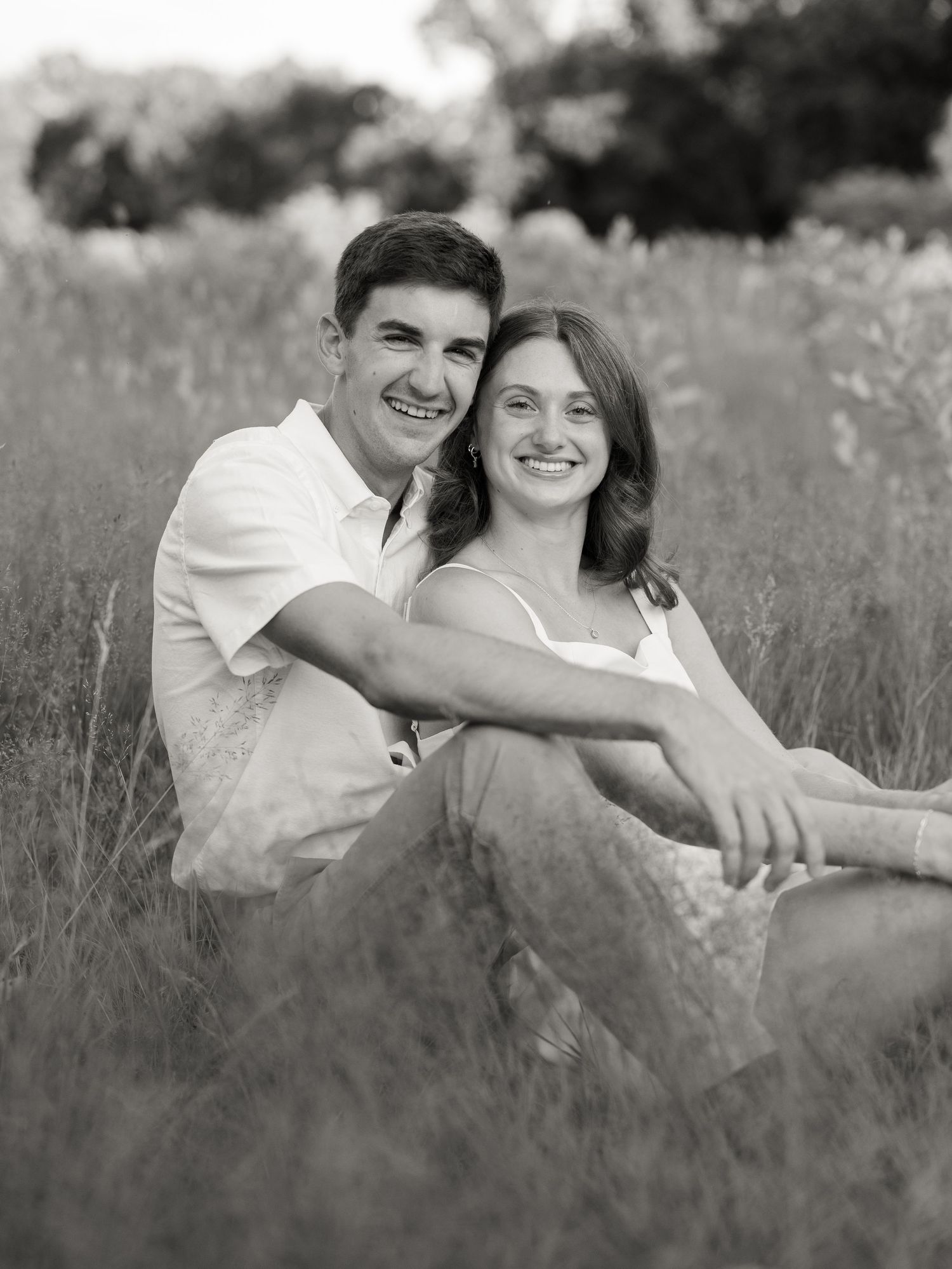 Black and white photo of a smiling couple sitting together in a field of tall grass outdoors.