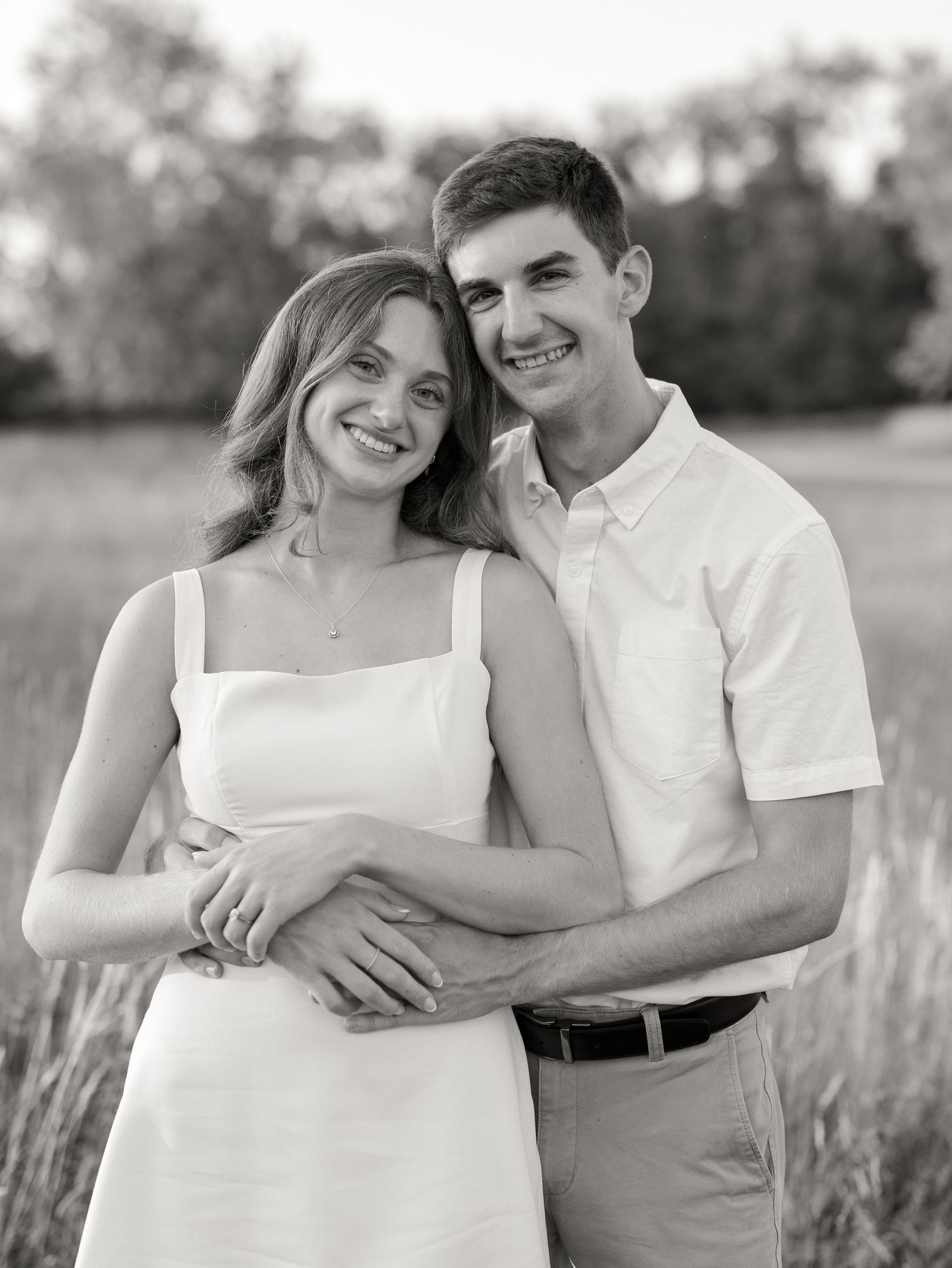 Black and white photo of a smiling young couple embracing outdoors in a field, woman in white dress.