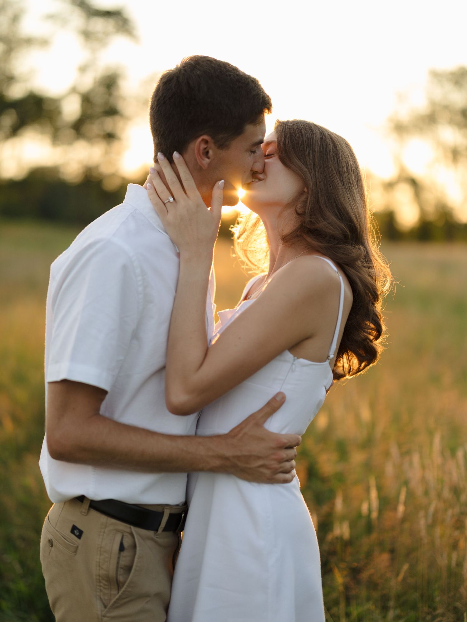 Couple sharing a romantic kiss in a golden sunset field, dressed in white and khaki, embracing tenderly.
