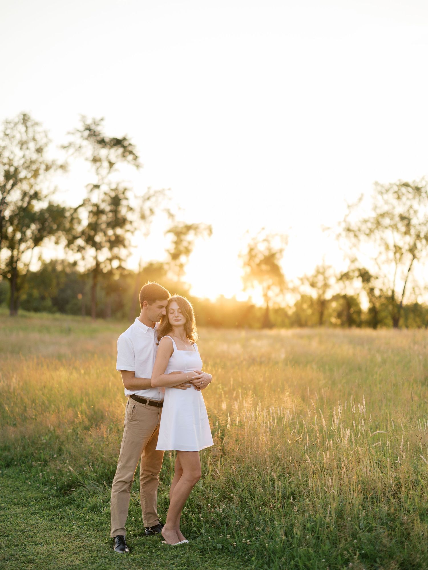 Couple embracing in golden sunset field, woman in white dress, man in khaki pants, surrounded by tall grass and trees.