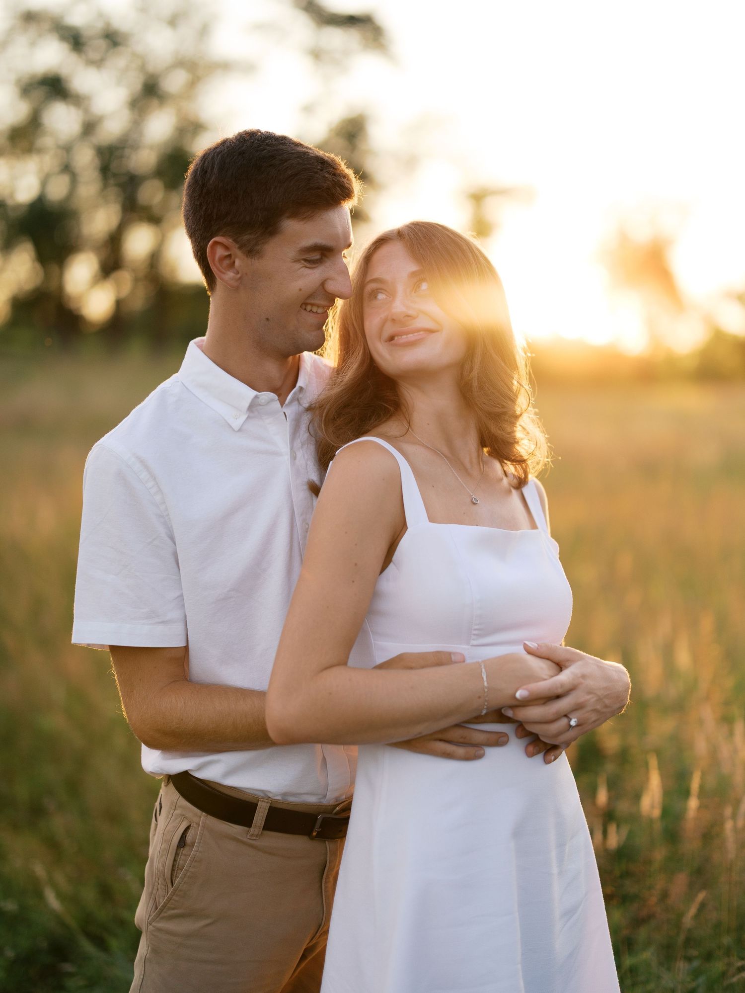 Couple embracing in golden sunset field, woman in white dress smiling, man in white shirt and khaki pants.