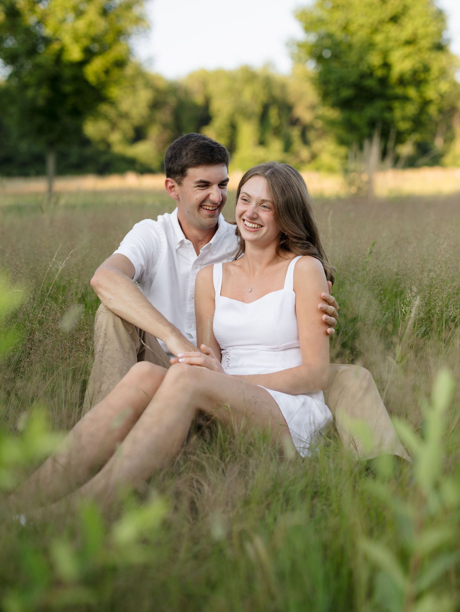 A smiling couple sits together in a lush green field, the woman in a white dress laughing joyfully.