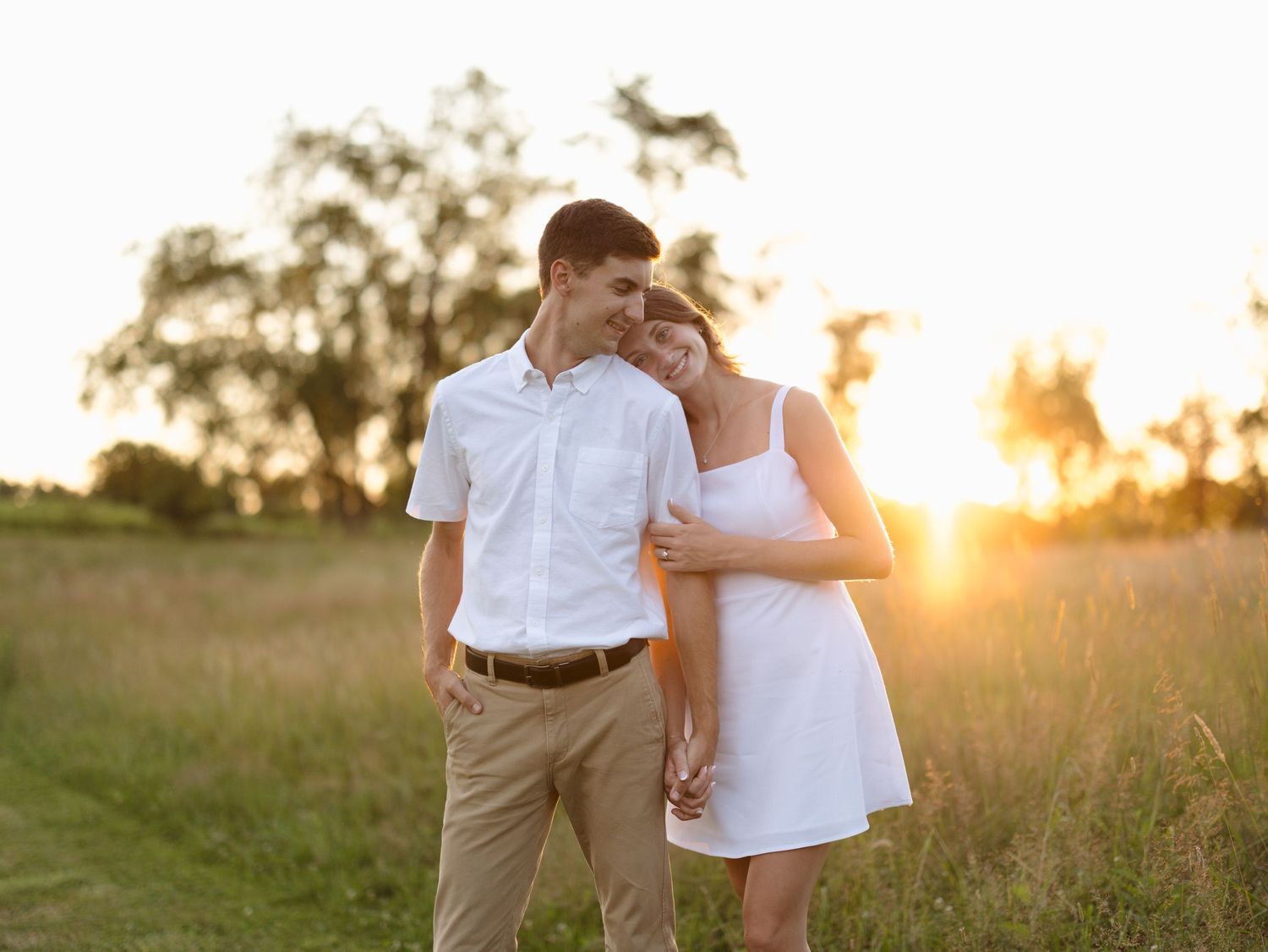 Couple in white outfits holding hands in a golden sunset field, sharing a romantic moment outdoors.