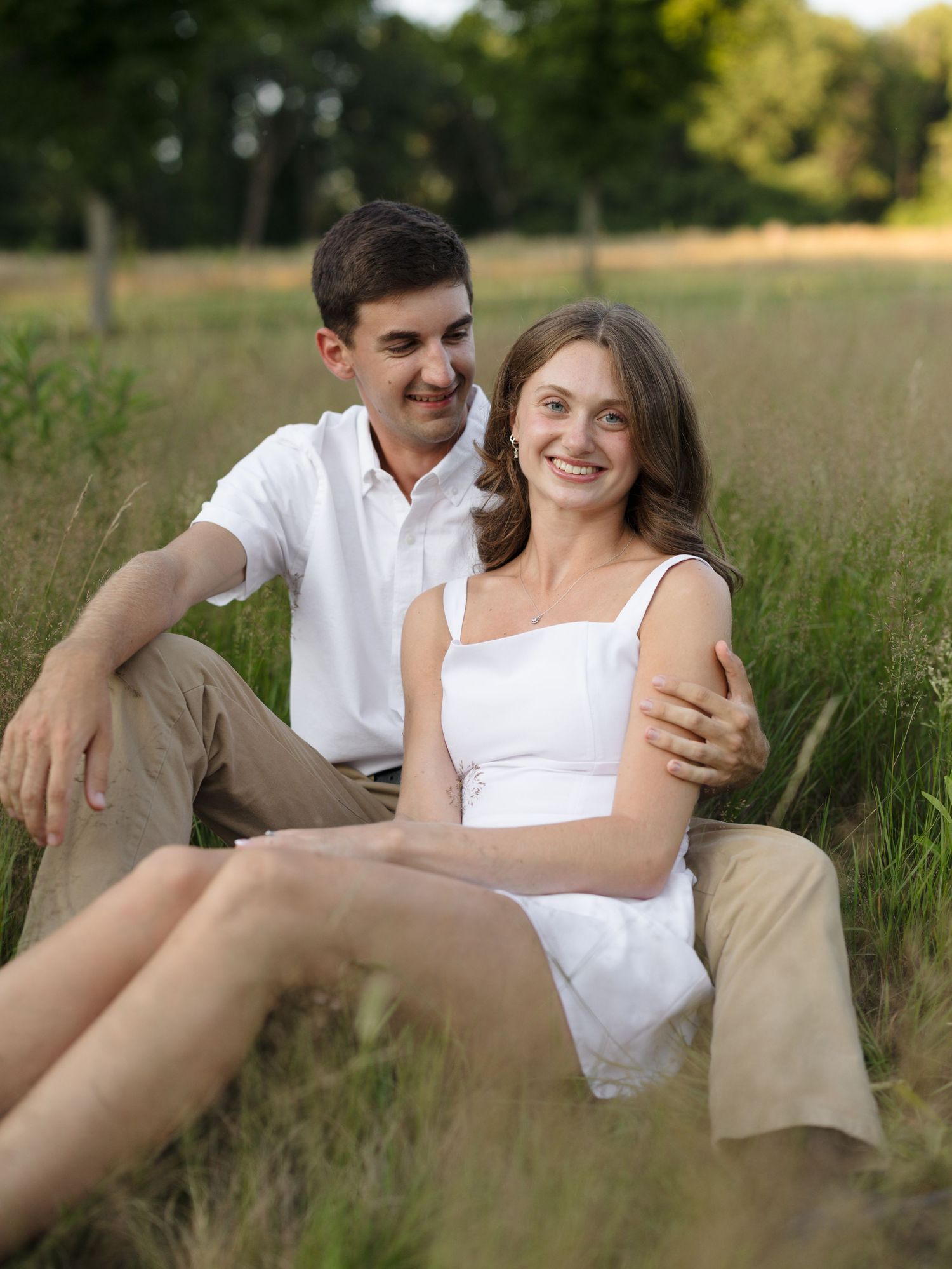 A young couple sitting together in a green meadow, smiling warmly, dressed in white tops and khaki pants.
