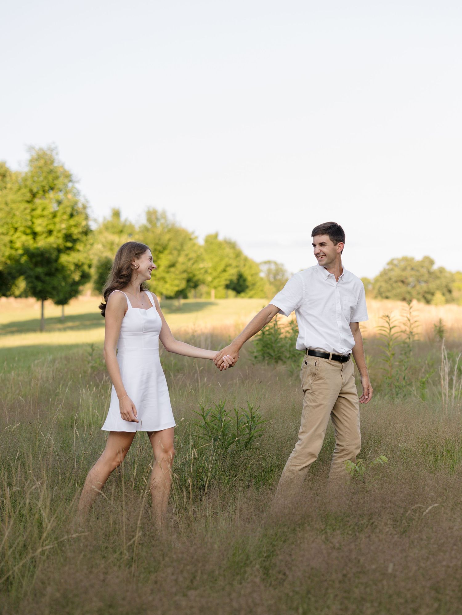 A couple holds hands and smiles while walking through a lush green meadow, woman in white dress, man in khakis.