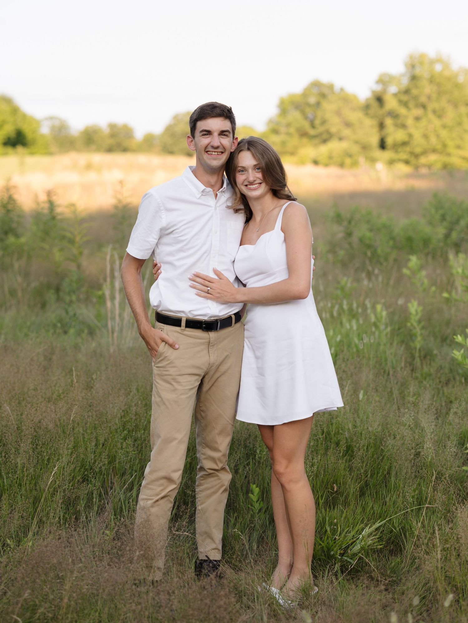 Couple posing together in a sunny field, man in white polo and khakis, woman in white mini dress, surrounded by green trees.