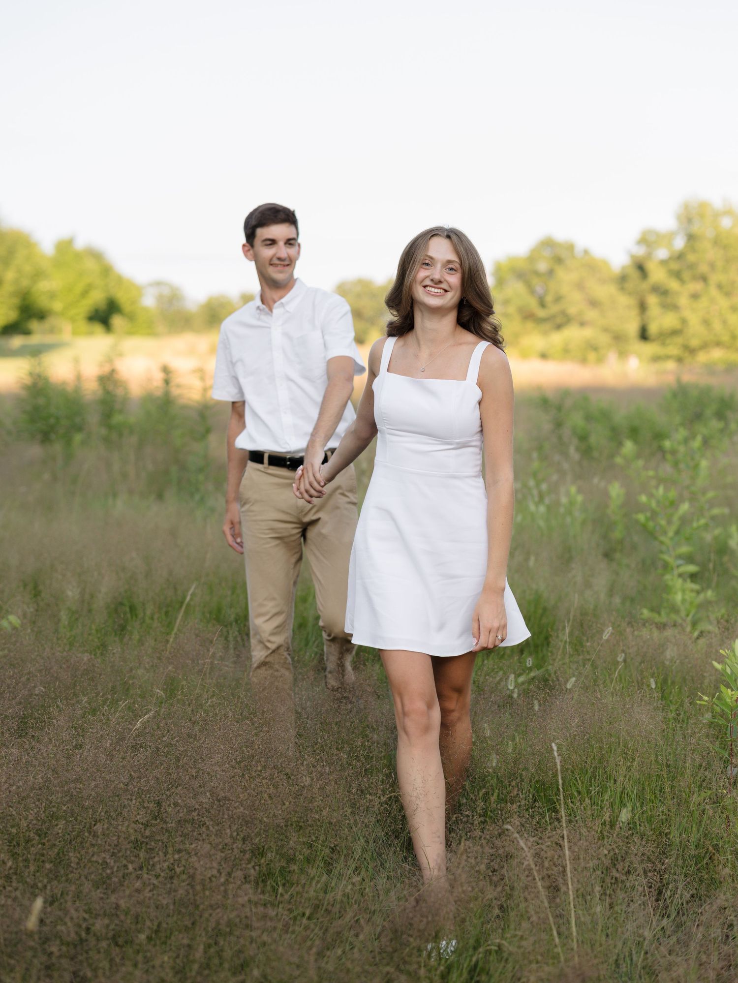 A couple holds hands walking through a lush green field, woman in white dress leads smiling, man in white shirt follows.