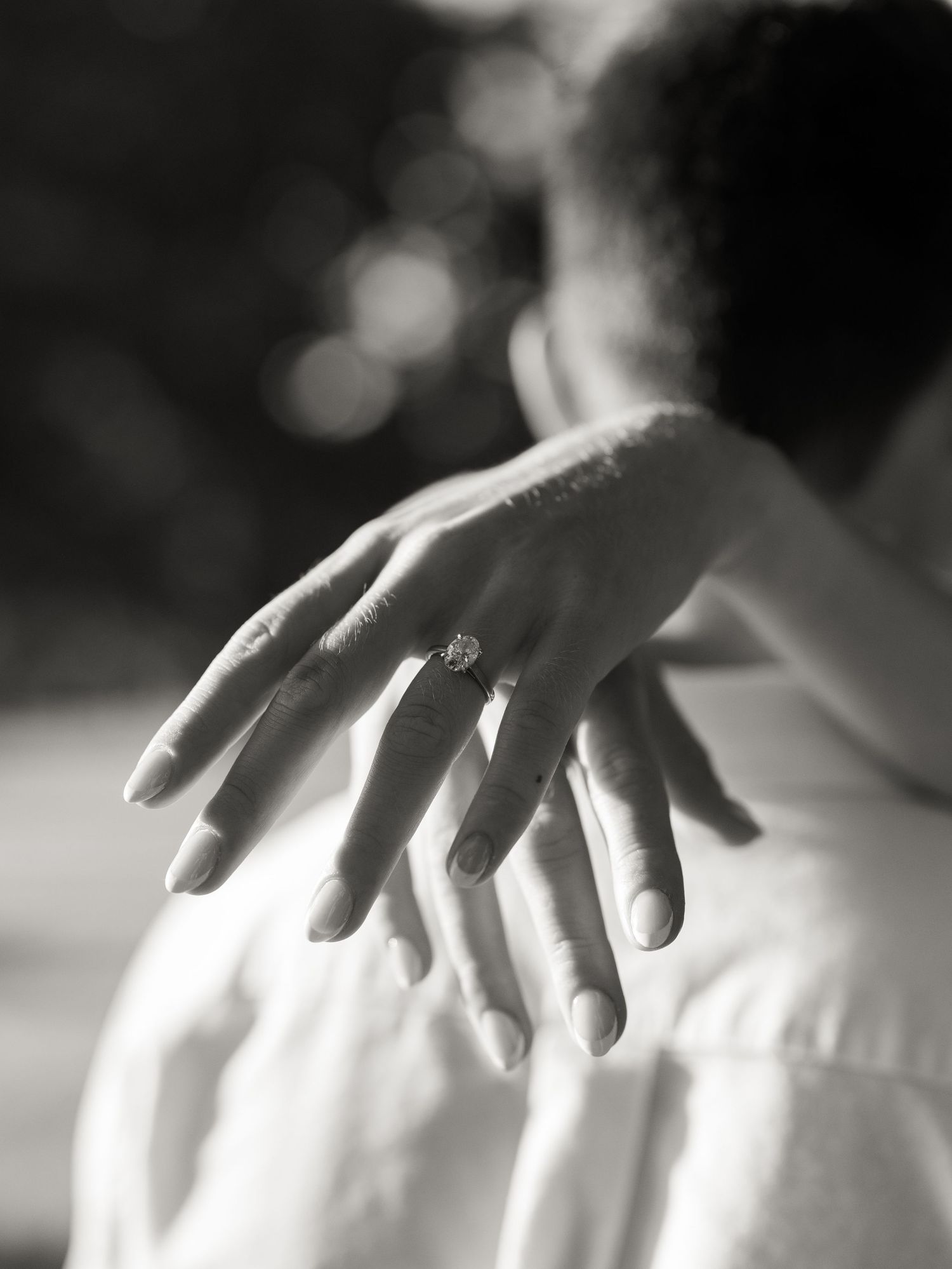 Close-up black and white photo of a woman's hand with a diamond engagement ring resting on a man's shoulder.
